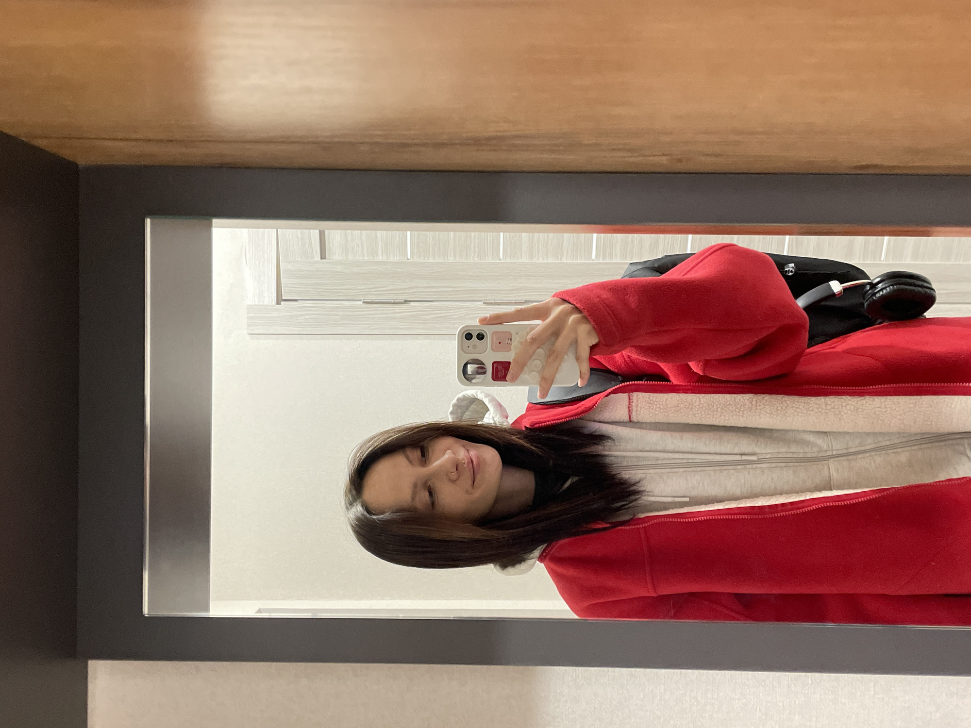A young woman is depicted taking a mirror selfie indoors, holding a white smartphone in her right hand. She has shoulder-length dark hair, a subtle smile, and a small nose piercing. She is dressed in a bright red fleece jacket worn over a light grey hooded top, which appears to have a soft, possibly sherpa lining.
The setting appears to be an indoor space, possibly a hallway or room, with a dark-framed rectangular mirror reflecting the scene. Behind her, in the reflection, are light-colored door or wall panels with distinct linear designs. Above the mirror, a wooden ceiling with a visible grain is seen. To the woman's right, also reflected in the mirror, are a black backpack and a pair of black over-ear headphones, suggesting an impending journey or recent arrival.
The smartphone case features small, colorful, square-shaped graphic elements, but no clearly legible text is visible. The lighting is even and consistent with an indoor environment. The image conveys a casual and relaxed mood. The context provided is Izhevsk, Russia.