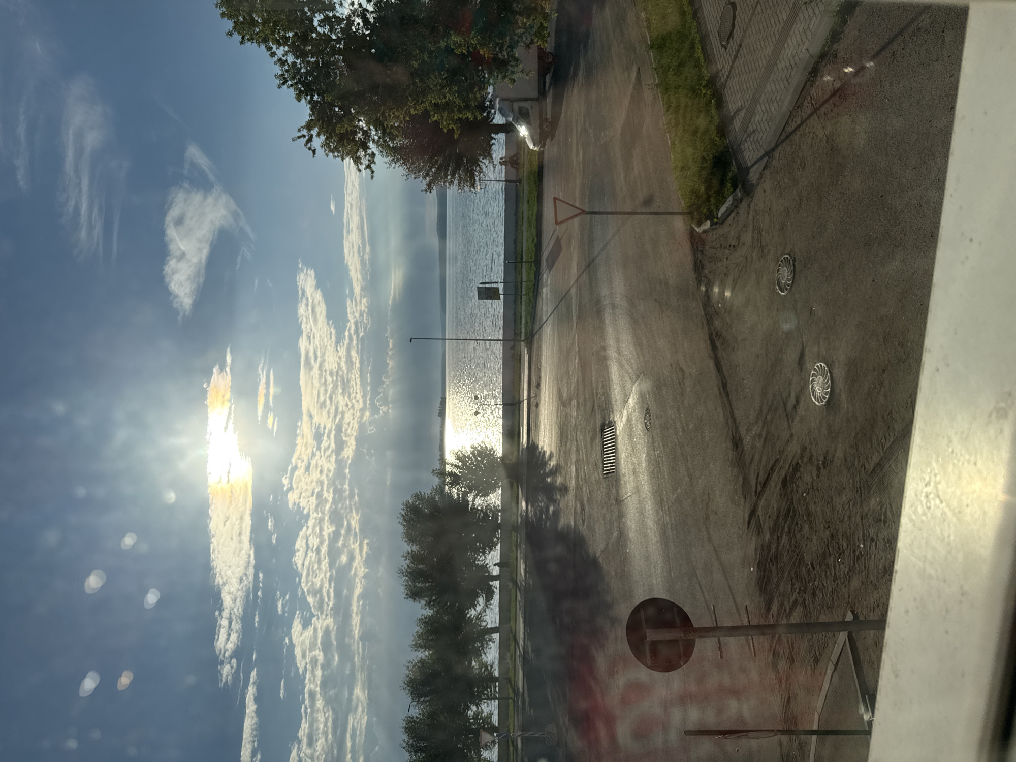 The image shows a view from a window of a body of water, likely a lake or river, bordered by a line of trees. The sun is bright, casting reflections on the water and creating sunbeams that filter through the clouds. In the foreground, there is a paved area with tire tracks and a few manhole covers. There are also road signs and a small white van parked on the right side of the frame. A single person is visible walking along the path by the water. The scene appears to be set in the city of Votkinsk, Russia, during daylight hours.
