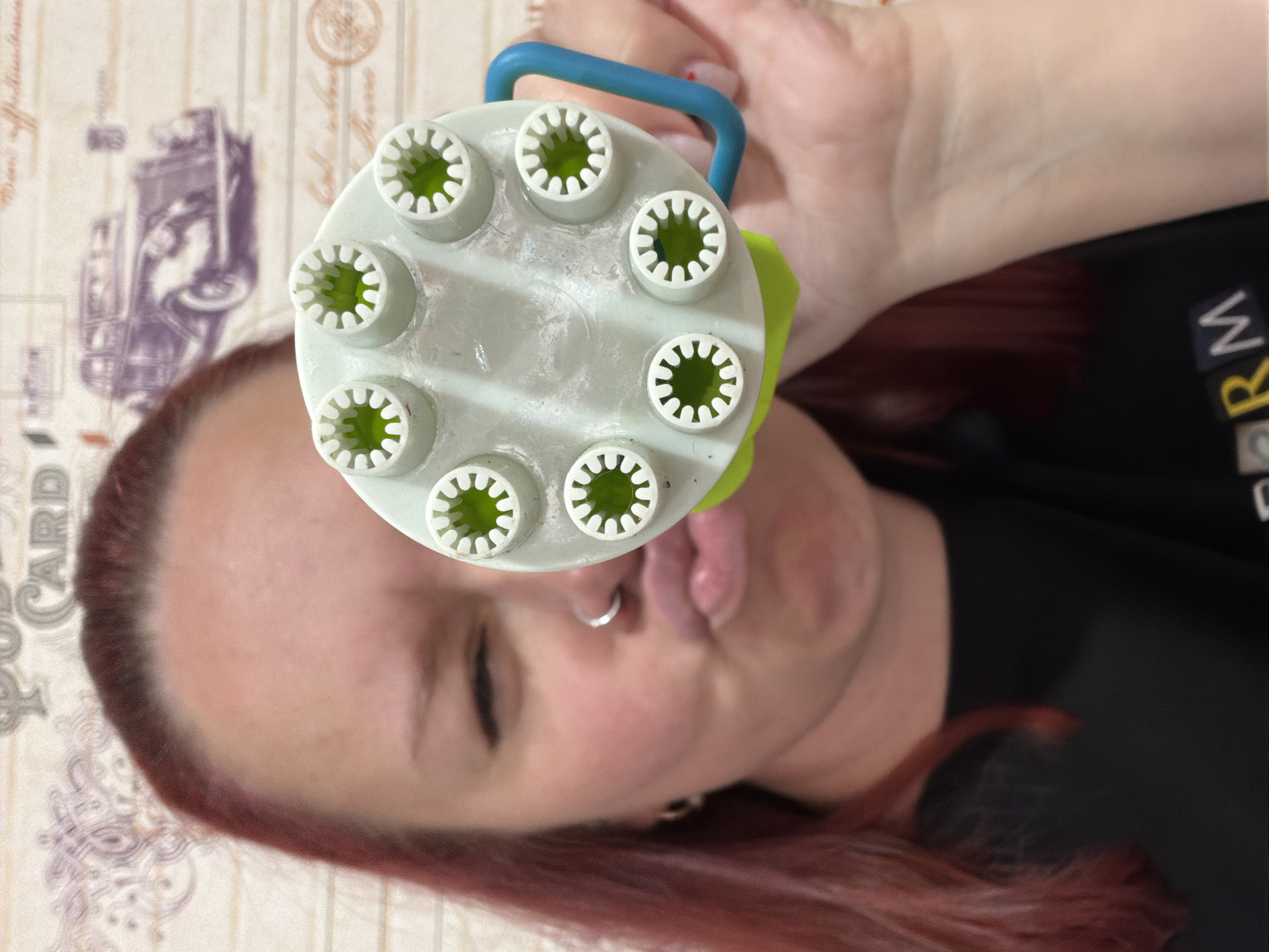 A person is holding a toy in front of their face. The toy appears to be a light green plastic device with seven circular openings containing a green liquid or gel. The toy has a blue handle. The person has fair skin, red hair, and is wearing a black shirt. The background is a patterned surface that resembles an old postcard. The location in the city of Chaikovsky, Russia is not discernible from the image.