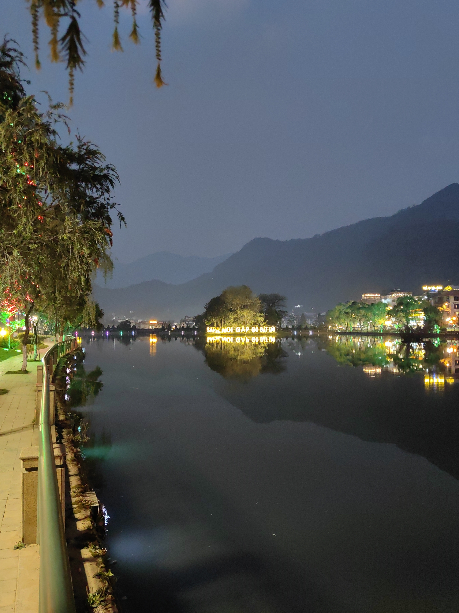 This nighttime image captures Sa Pa Lake in Sa Pa, Vietnam. In the foreground on the left, a paved pedestrian walkway is bordered by a dark metal railing. Trees alongside the path are decorated with multi-colored string lights, including red, green, and yellow, with additional yellow hanging ornaments visible from an overhead branch. The dark, calm surface of the lake occupies the central and lower portions of the frame, extensively reflecting the artificial lights. A small, treed island is positioned centrally in the lake, displaying large, illuminated yellow letters that spell "NƠI GẶP GỠ BẤT TÂN," which are clearly reflected in the water below. Across the lake, on the far bank, multiple buildings are visible with illuminated windows and facades, interspersed with trees that are also lit, primarily with green lights. A bridge-like structure with several arches and lights spans a section of the far bank. Dark, indistinct mountain ranges rise in the background under an overcast or dusky blue sky. No individuals or active subjects are present in the scene.
