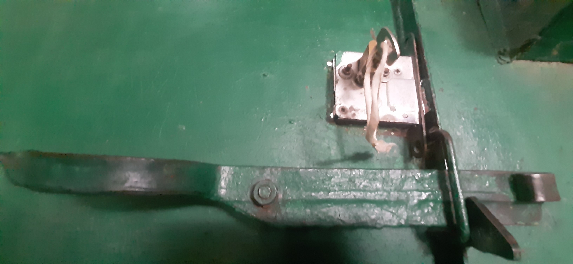 The image shows a close-up of a green, weathered surface, possibly a metal door or wall, with a tarnished metal latch mechanism. The latch consists of a long, horizontal bar with a tapered end, secured by a bolt. Attached to this bar is a pivoting lever mechanism with a strap or wire hanging from it. The surface is textured and shows signs of wear, with scratches and some flaking paint. The environment appears to be industrial or utilitarian, with no visible context to suggest a specific location in Yessentukskaya, Russia. There are no people or animals present in the image.