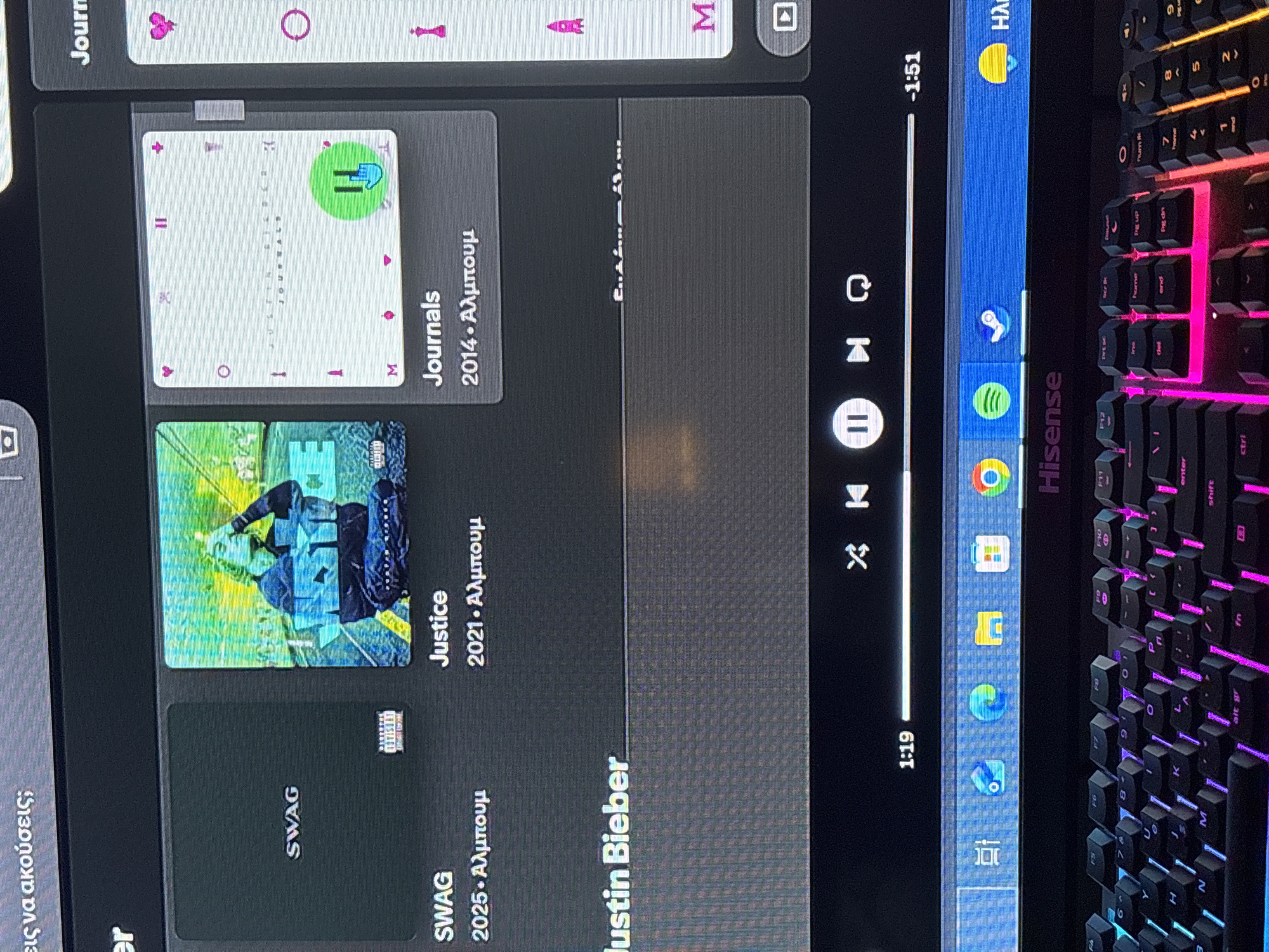 The image shows a computer screen displaying the Spotify application. The application is in the process of playing music by Justin Bieber. The albums "SWAG", "Justice", and "Journals" are visible as recently played or available music in the queue. The song is currently playing and is at the 1 minute 19 second mark, with 1 minute 51 seconds remaining. To the right of the screen is part of a keyboard, with RGB lighting. The location is most likely at a computer desk in a home or office environment. There is no information to connect the image to the city of Kallithea, Greece.