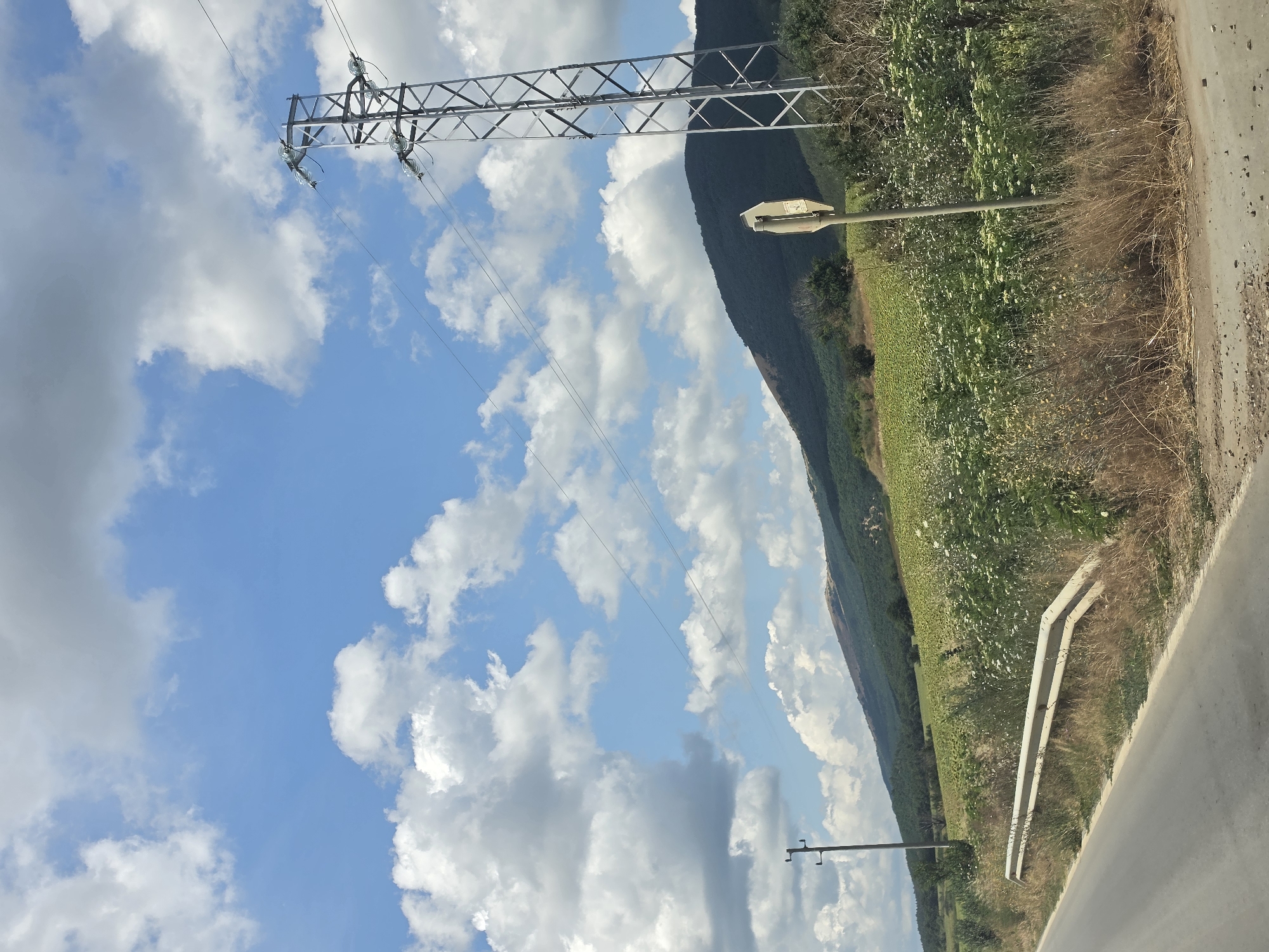 A road cuts through a rural landscape under a partly cloudy blue sky. A metal guardrail runs along the side of the asphalt road, bordering dry grass and vegetation. Beyond the roadside, a field of green crops, possibly sunflowers, stretches towards a rolling, tree-covered hill. In the background, a large, silver metal electricity pylon stands tall, with power lines extending diagonally across the sky. A simple wooden utility pole is visible further to the left. A signpost with a damaged white sign is positioned near the electricity pylon. The image appears to be taken on a sunny day.
