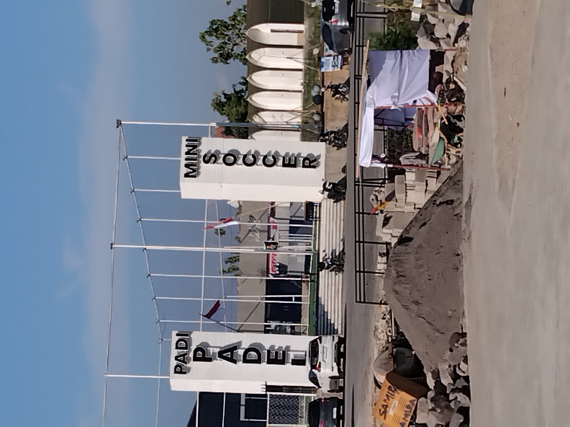 The image captures a daytime exterior scene in Papahan, Indonesia, featuring a facility with prominent signage for "PADI PADELF" and "MINI SOCCER". The "PADI PADELF" sign is displayed vertically on a white pillar, and "MINI SOCCER" is also on a white pillar, with "SOCCER" written vertically. In the background, several arched structures are visible, possibly part of the facility's design. In front of the building, there is a set of outdoor stairs, and several motorcycles are parked or being ridden by individuals. Two cars are also visible on the road in front of the facility. The foreground shows a construction or renovation site with piles of sand and gravel, building materials like concrete blocks, a wheelbarrow, and a yellow construction equipment. A white tent structure is also present in the foreground. The sky is clear and blue.