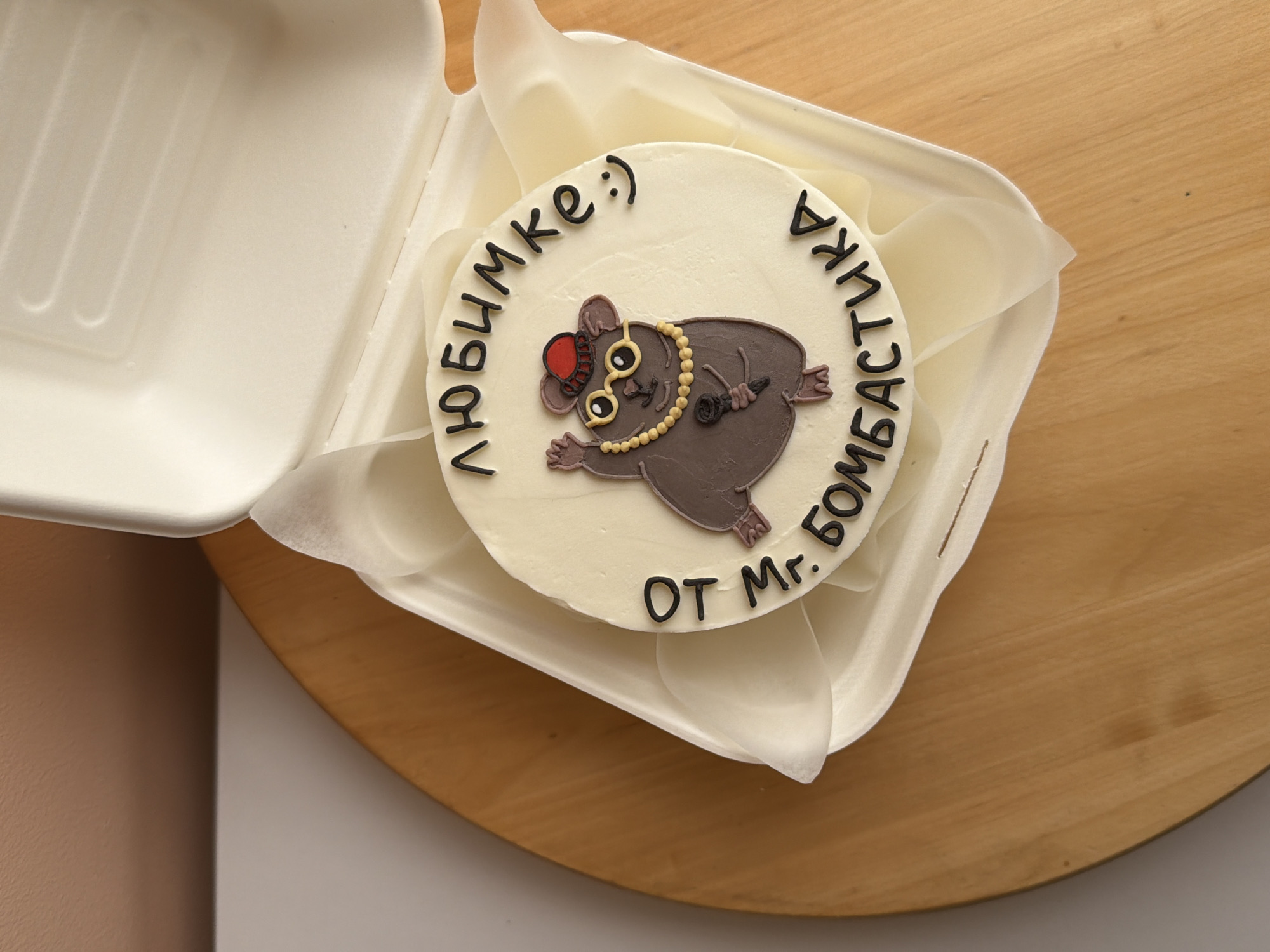 A top-down shot captures a small, round cake nestled inside a white, disposable take-out container. The cake is covered in smooth, off-white frosting and features a whimsical illustration of a mole. This cartoon-like mole is depicted lying on its back, wearing a red fedora-style hat, a gold monocle, and a long, golden beaded necklace. It holds a black microphone in one paw, suggesting a performance or playful persona.
Around the upper curve of the cake, black text in Cyrillic script reads "ЛЮБИМКЕ :)" which translates to "To my darling/sweetheart :)". Below the mole, on the lower curve of the cake, more black text reads "ОТ Mr. БОМБАСТИКА", meaning "From Mr. Bombastic". The cake is lined with a translucent, white parchment-like paper within the container.
The container rests on a light-colored wooden surface, possibly a cutting board or table. To the left, a portion of a light brown or beige wall or vertical surface is visible, while a white surface appears in the lower left background. The overall impression is one of a personalized gift, likely for a celebration or special occasion, presented indoors.