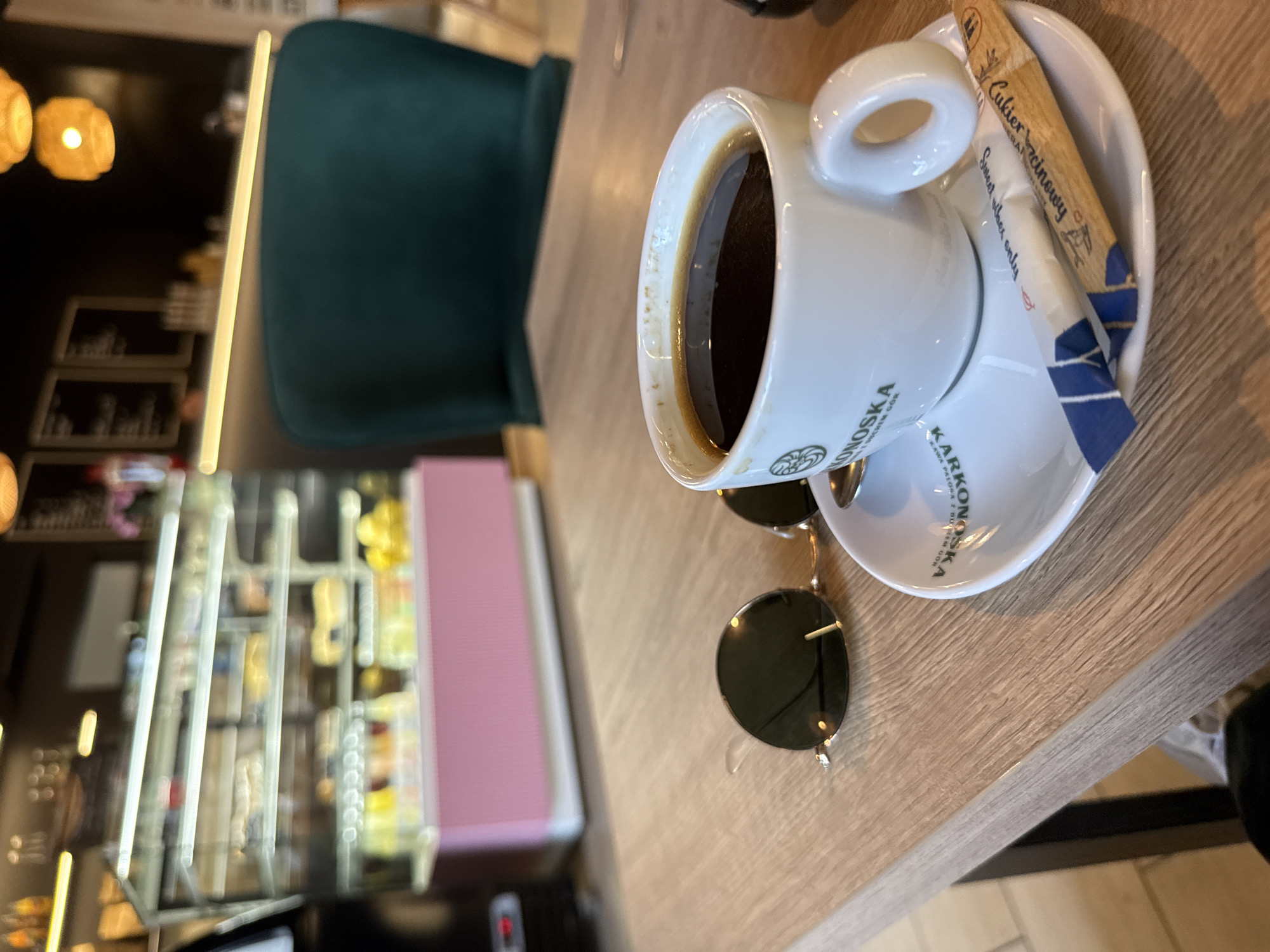 The image shows a close-up view of a white cup of black coffee with a sugar packet and a pair of sunglasses resting on a wooden table. The coffee cup has text on it which is partially visible. A sugar packet with text like "Cukier Brzozowy" and "Sweet vibes only" is placed on the saucer. The sunglasses are round with dark lenses. In the background, out of focus, is a dark green chair and shelves displaying various items. The scene appears to be set in a cafe or restaurant. The lighting suggests it could be daytime. The presence of coffee and sunglasses indicates a casual setting. Some text on the cup seems to be in Polish, mentioning "KRONOSKA" and "KAWA Z MLEKIEM". The overall impression is of a relaxed moment, possibly a break or a pause during the day. The location is identified as Szklarska Poręba, Poland.