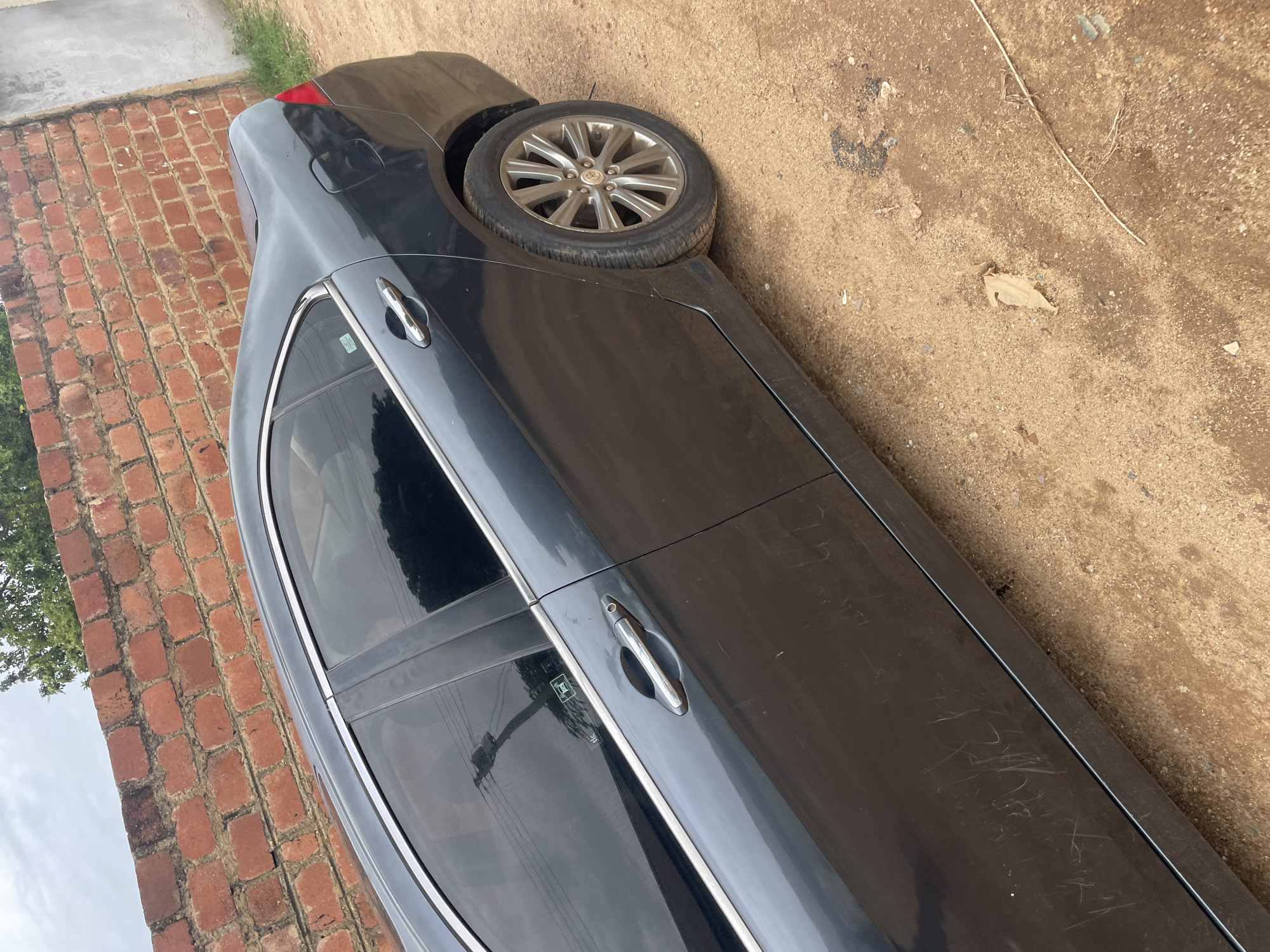 A dark gray sedan is parked on a dirt road next to a red brick wall in an outdoor setting. The car is viewed from the side, with the rear passenger door, rear wheel, and part of the rear bumper visible. The car's windows are tinted. A small, light-colored sticker with green text and a logo is present on the rear passenger window. The ground is a mixture of sand and small stones. A few dried leaves and twigs are scattered on the ground. The brick wall appears weathered and is partially covered by greenery at the top. The sky is overcast. The overall scene suggests an informal parking area.
