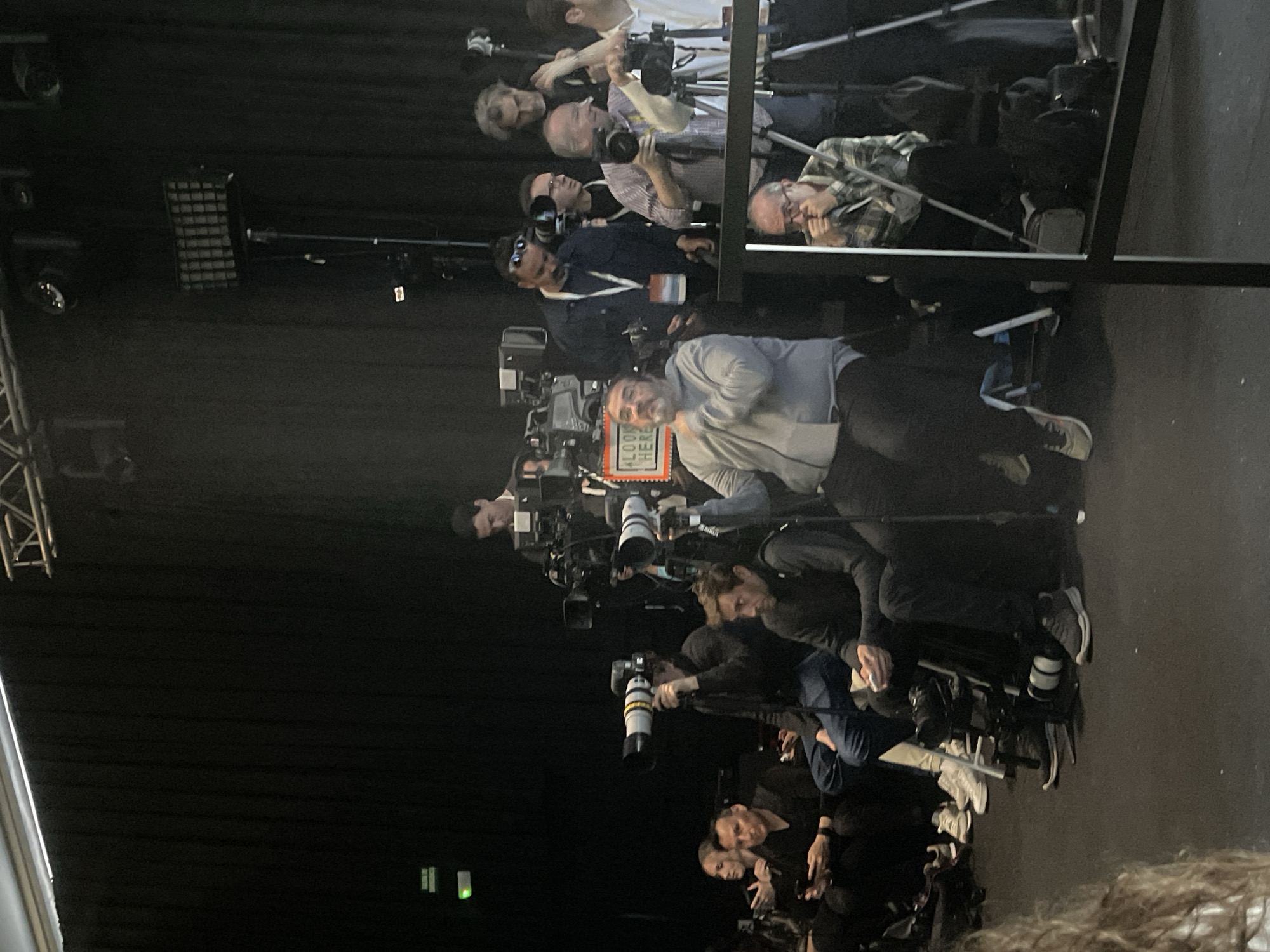 The image shows a group of photographers, mostly men, positioned in a line, likely behind a barrier or railing, facing forward. They are equipped with professional-grade cameras, some mounted on tripods, and long telephoto lenses. The lighting is bright, suggesting a stage or event setting. A sign with orange letters is visible on one of the cameras. The backdrop is dark, possibly a curtain or wall. The image does not provide information about Barcelona, Spain, so I cannot comment on the location.