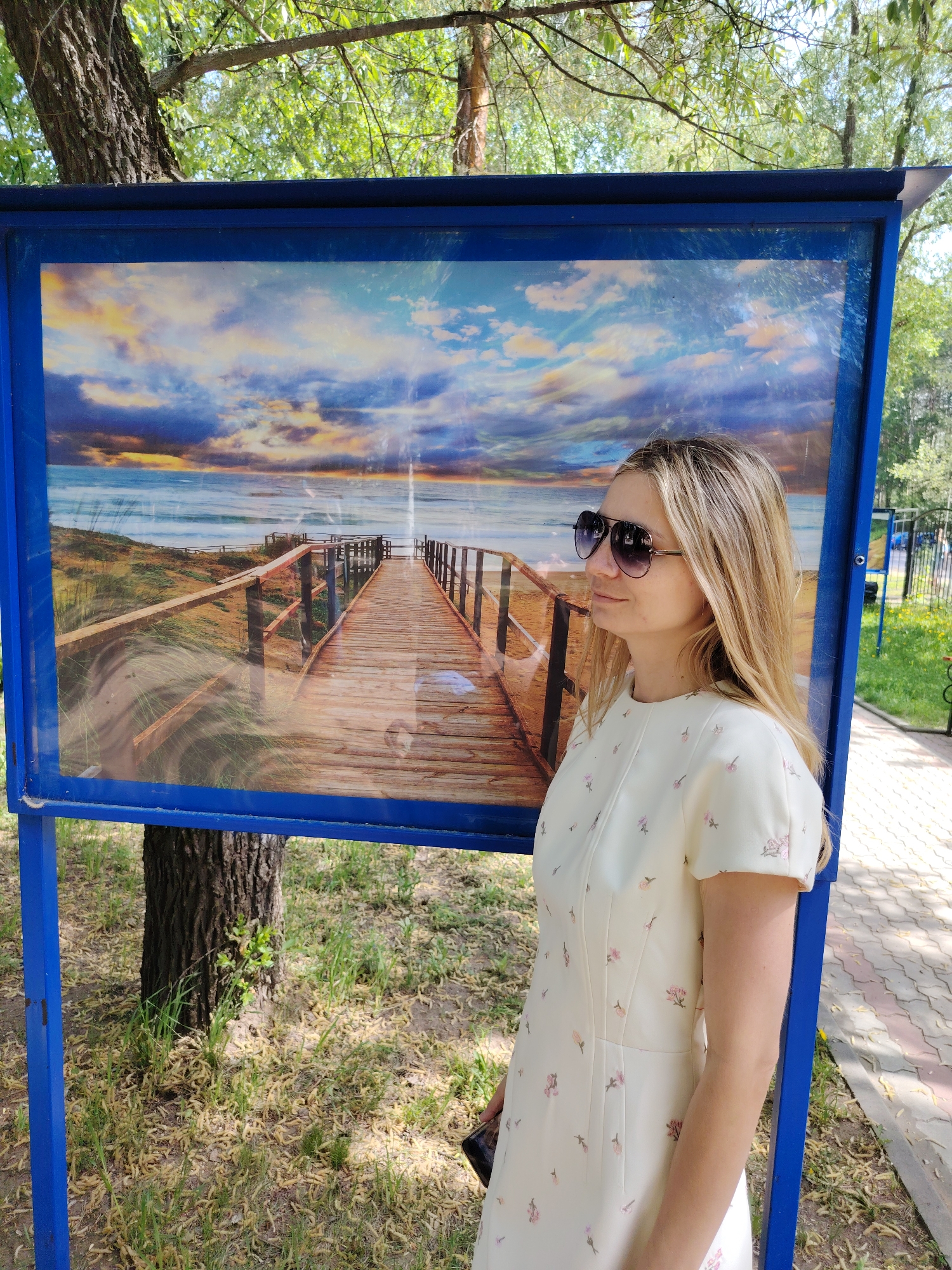 A woman in a light-colored dress and sunglasses stands next to a large blue display board in what appears to be a park or outdoor setting. The display board contains a picture of a wooden boardwalk leading towards the ocean under a dramatic sunset sky. The woman is positioned to the right of the board and is looking to her right. The background includes trees, grass, and a brick-paved walkway. This is in the city of Chaikovsky, Russia.