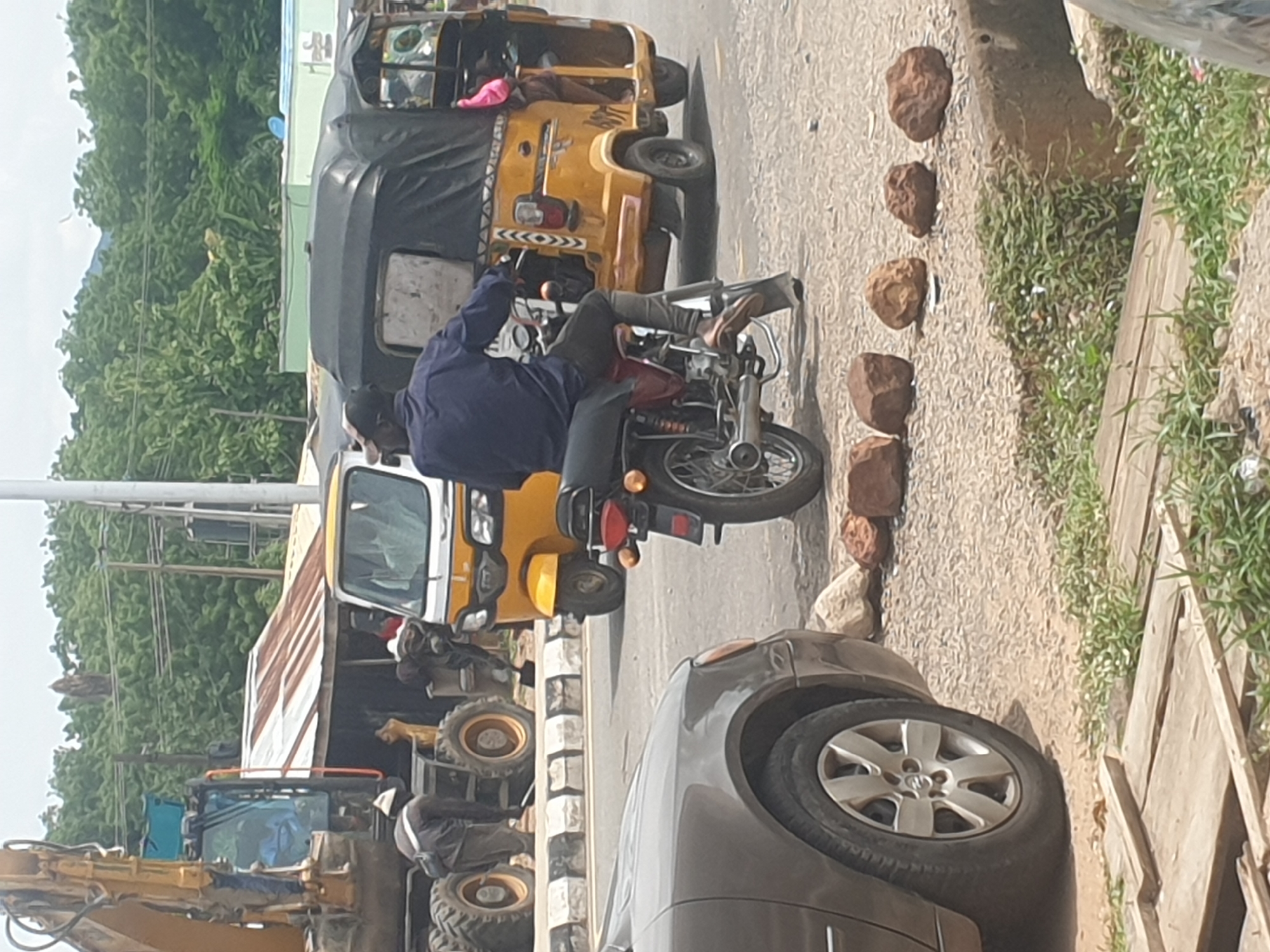 The image shows a street scene in Adankolo, Nigeria, with various vehicles and objects present. A man on a motorcycle is in the foreground, wearing a blue shirt and dark pants. Next to him is a gray car, with part of the front wheel visible. Behind the motorcycle, there are two yellow three-wheeled taxis (Keke Napep) and an earthmover. Five rocks are lined up along the right edge of the road next to vegetation and wooden planks. The image is rotated.
