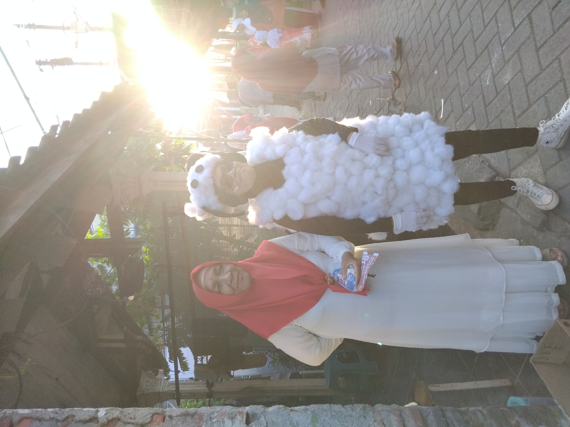 In the city of Kepuh Kiriman, Indonesia, two individuals are positioned on a cobblestone pathway under a weathered wooden overhang. The person on the left, a woman, wears a long, cream-colored dress and a vibrant red hijab. She holds a bottle of water and a small brochure in her hand. To her right, another individual is dressed in a costume resembling a sheep, complete with a white fluffy body, a black and white sheep-head accessory, white gloves, and sunglasses. They are also wearing a flag sticker on their cheek. The background reveals a street scene with other people, some in traditional attire, and sunlight streams in from the right, creating lens flare and illuminating the scene. The environment suggests an outdoor event or gathering.
