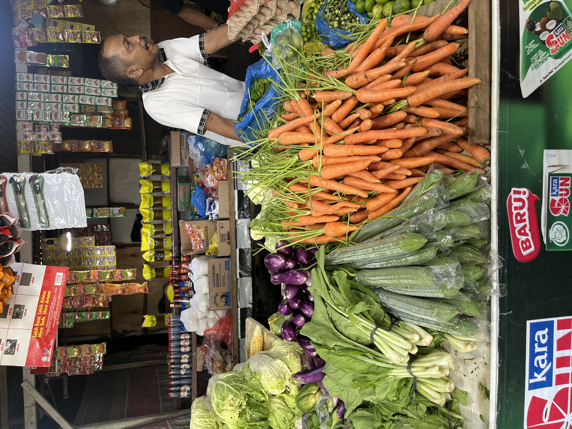 The image depicts a market stall in Lhokseumawe, Indonesia, with a variety of fresh produce. A middle-aged man with short, dark hair and a mustache, wearing a white polo shirt with a black and white striped collar, stands behind the stall, looking to his right. He appears to be a vendor. The stall is laden with vegetables, including large heads of cabbage, bunches of leafy greens, bundles of carrots, eggplants, and long, green gourds. Behind the vegetables, there are shelves stocked with various packaged goods, including bottles of sauces, packets of spices, and bags of flour. In the background, more shelves are visible, filled with a wider assortment of products. The market appears to be covered by a corrugated metal roof. The overall scene is one of a bustling local market, showcasing the availability of fresh ingredients.
