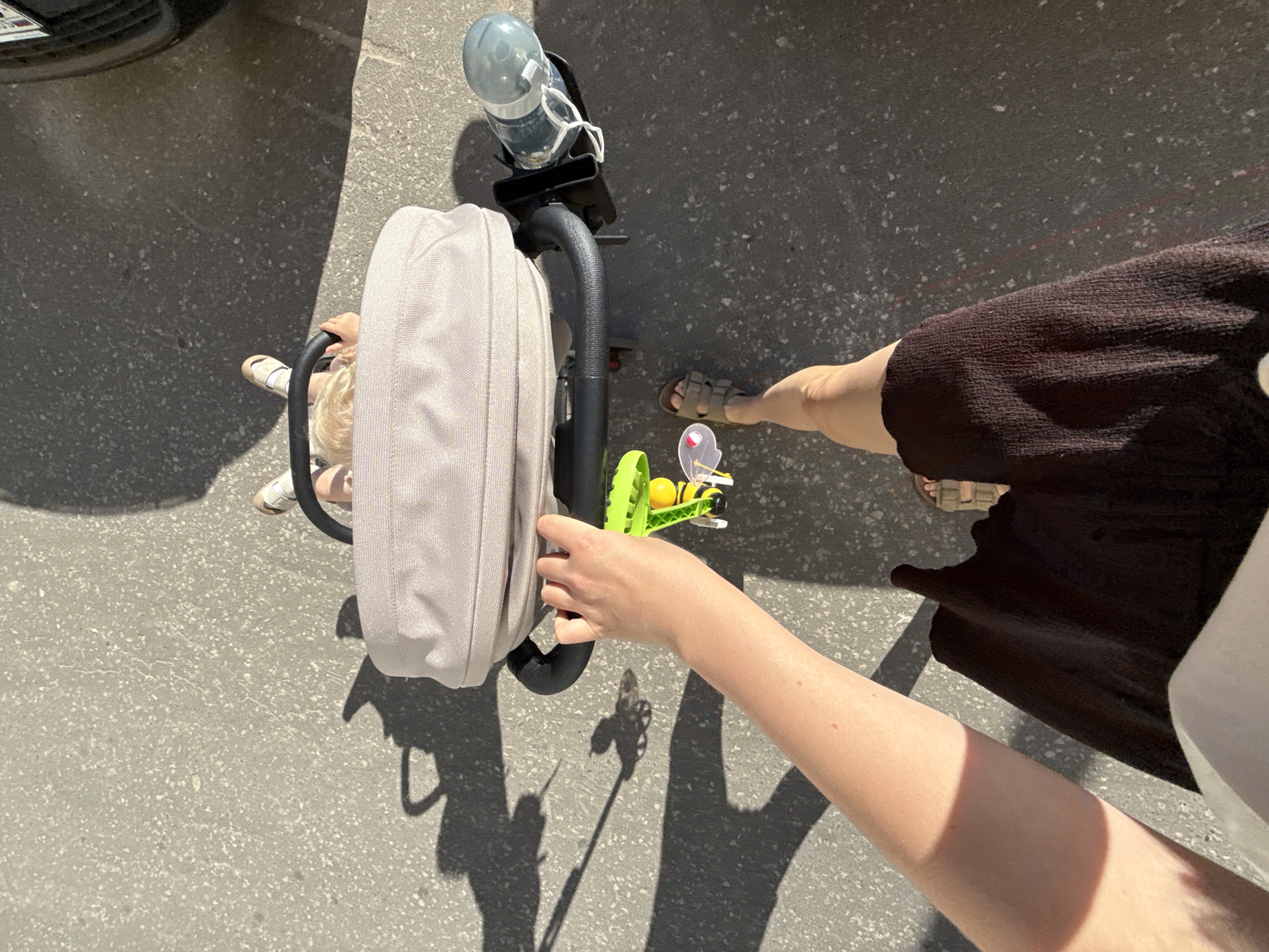 A high-angle shot shows a person pushing a stroller on a sunny day. The scene is set on an asphalt surface, possibly a sidewalk or street. A portion of a car is visible in the upper left corner. The stroller has a light gray bag attached to it and a plastic bottle holder with a water bottle. A bright green toy with a yellow bee is clipped to the stroller's handle.
Two people's legs are visible. One set of legs, belonging to a child with blonde hair, is positioned near the stroller's frame, wearing white sandals. The other set of legs, belonging to an adult, is wearing brown sandals and a dark brown skirt. The adult's arm reaches out, touching the toy on the stroller. The sunlight casts strong shadows, indicating it is daytime. The location is identified as Vladimir, Russia. No visible text is present in the image.