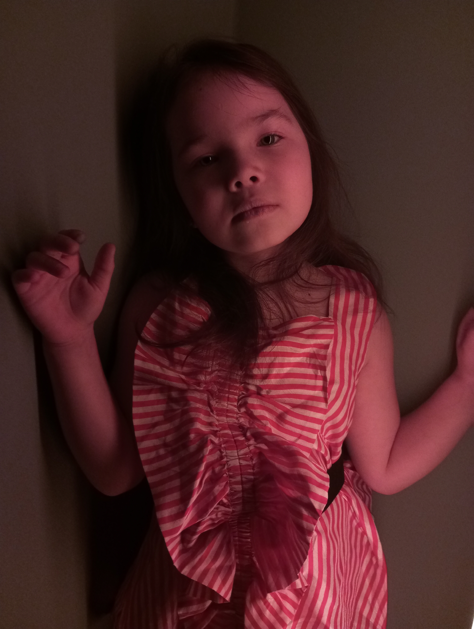 A young girl with dark hair is pressed against a dark-colored wall, possibly in a corner. She is wearing a red and white striped dress. The lighting in the image is red. There is no information to determine the location to be in the city of Pervomaysky, Russia.