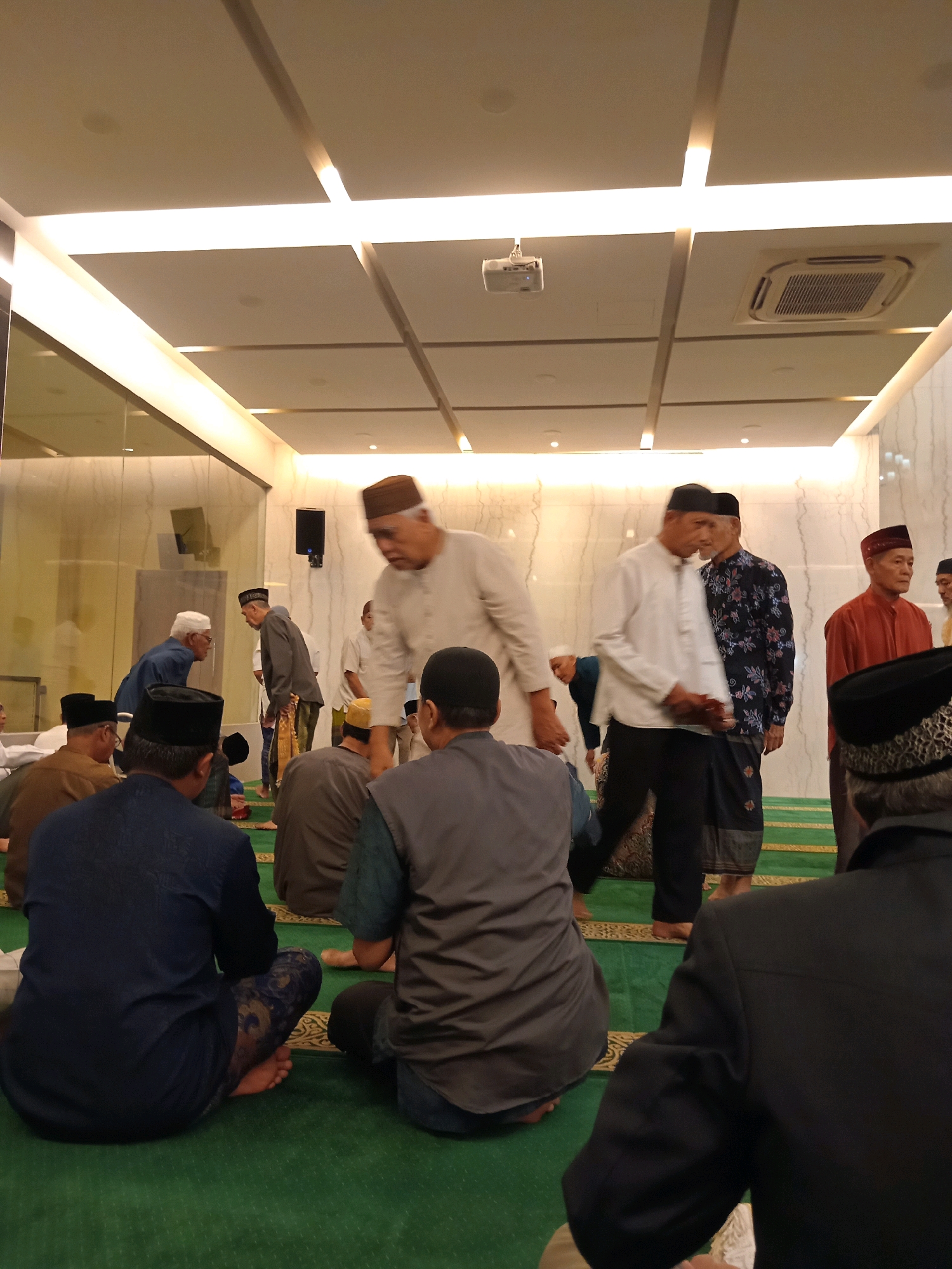 This image depicts a gathering of Muslim men indoors, likely at a mosque or community hall in Randuagung, Indonesia, for prayer or a religious event. The setting appears modern with a drop ceiling featuring integrated lighting and an air conditioning unit. A projector is mounted on the ceiling.
Several individuals are visible, mostly from the back or side, sitting or standing on a green carpet with patterned prayer mats. Many are wearing traditional Islamic attire, including peci (caps) and sarongs or long shirts.
In the foreground, on the left, a man in a dark blue shirt and patterned pants is seated, facing away from the camera. To his right, another man in a grey vest over a blue shirt is also seated. In the center of the image, a man in a light-colored long-sleeved shirt and dark pants is standing and appears to be interacting with another man to his left, who is wearing a dark patterned shirt and sarong. Further to the right, a man in a red shirt and cap is standing and looking towards the left. Other individuals are scattered throughout the scene, some with their backs to the camera, and some in the background interacting or preparing for prayer. The atmosphere suggests a communal religious observance.