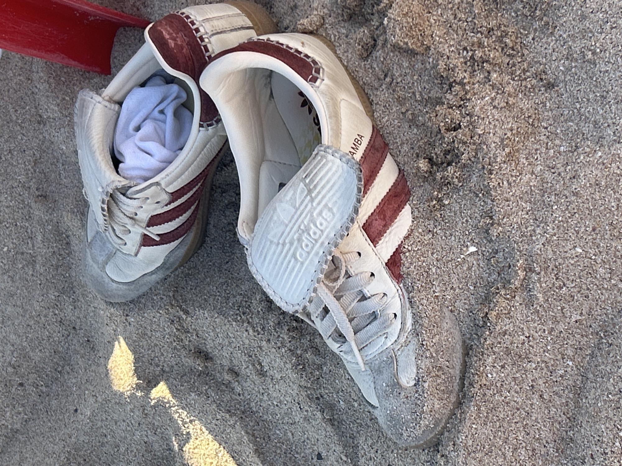 The image shows a pair of Adidas Samba shoes on a sandy surface. One of the shoes has a white sock stuffed inside. The shoes are off-white with dark red stripes. The sand is light brown. The location is unspecified, but the prompt suggests it's in Mazatlán, Mexico. No people are visible.