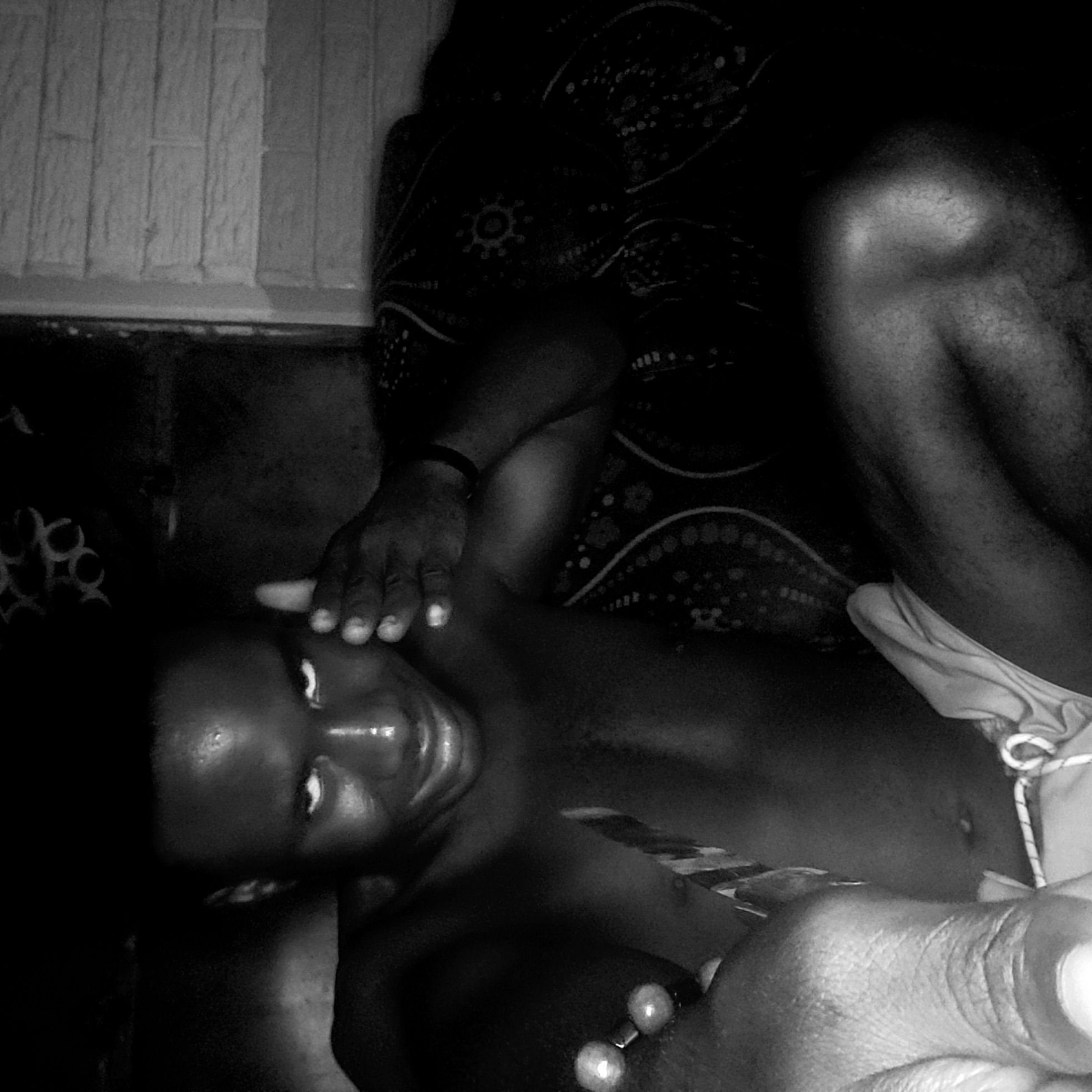 A person with dark skin is lying on a surface in Narayi, Nigeria. Another person is seated nearby. A hand rests on the reclining person’s forehead. A patterned cloth is near the person's head. A section of a wall with a rectangular pattern is visible. The reclining person is wearing what appears to be a beaded bracelet.