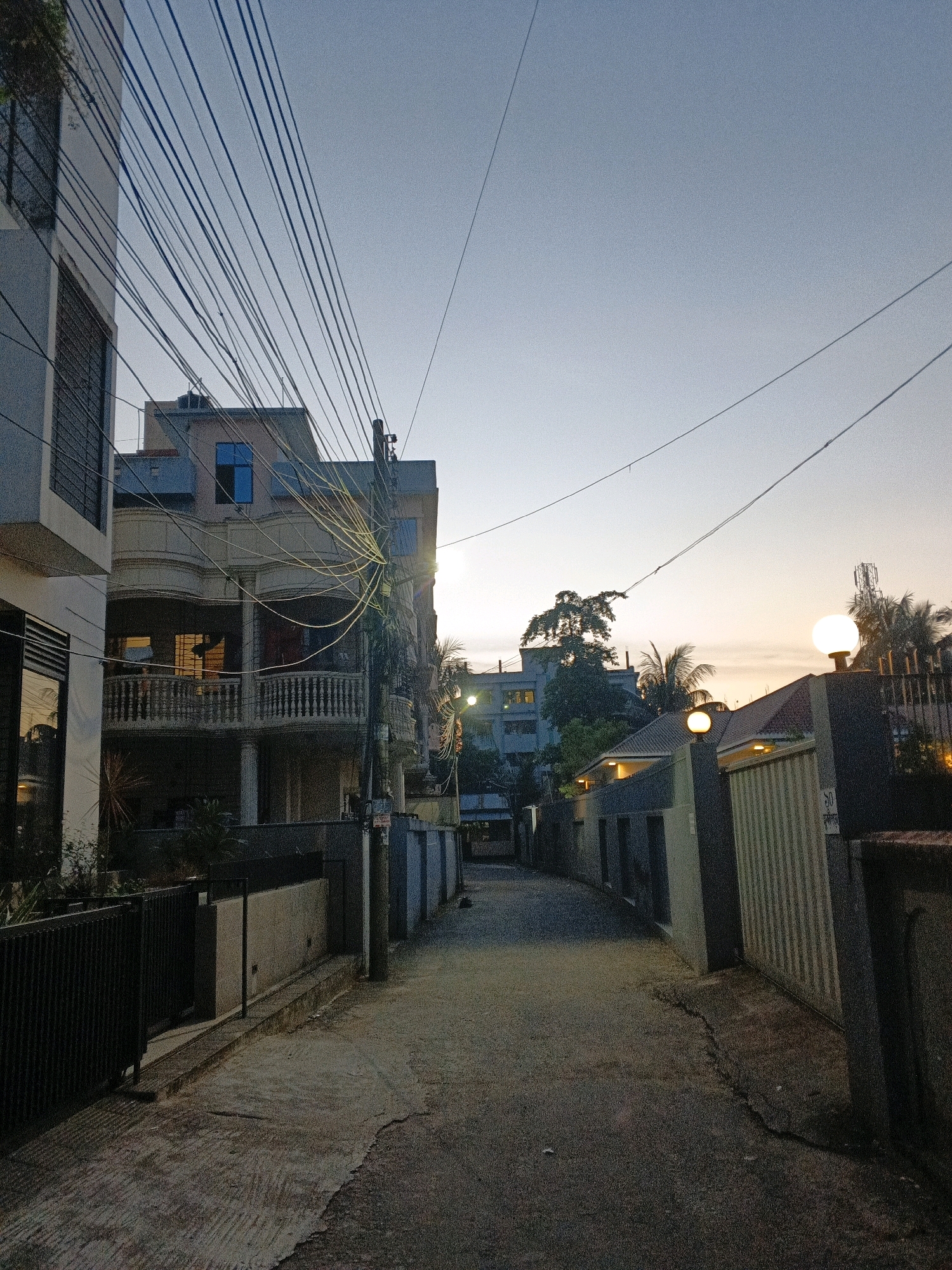 A narrow, paved alleyway runs between buildings in Sylhet, Bangladesh, during twilight. Overhead, a dense network of black electrical wires crisscrosses the sky, obscuring portions of the buildings and the pale, gradient sky. The buildings on either side of the alley are a mix of modern and older architecture. On the left, a sleek, multi-story building with dark-tinted windows is visible. To the right of that, a building with a cream-colored facade features balconies with ornate railings and lit windows, suggesting interior activity. Further down the alley, the sky is tinged with orange and yellow from the setting sun, illuminating palm trees and a telecommunications tower in the distance. Several outdoor light fixtures, some circular and some lamp-like, are illuminated, casting a warm glow. The alley pavement is uneven and cracked in places. No people are visible in the scene.