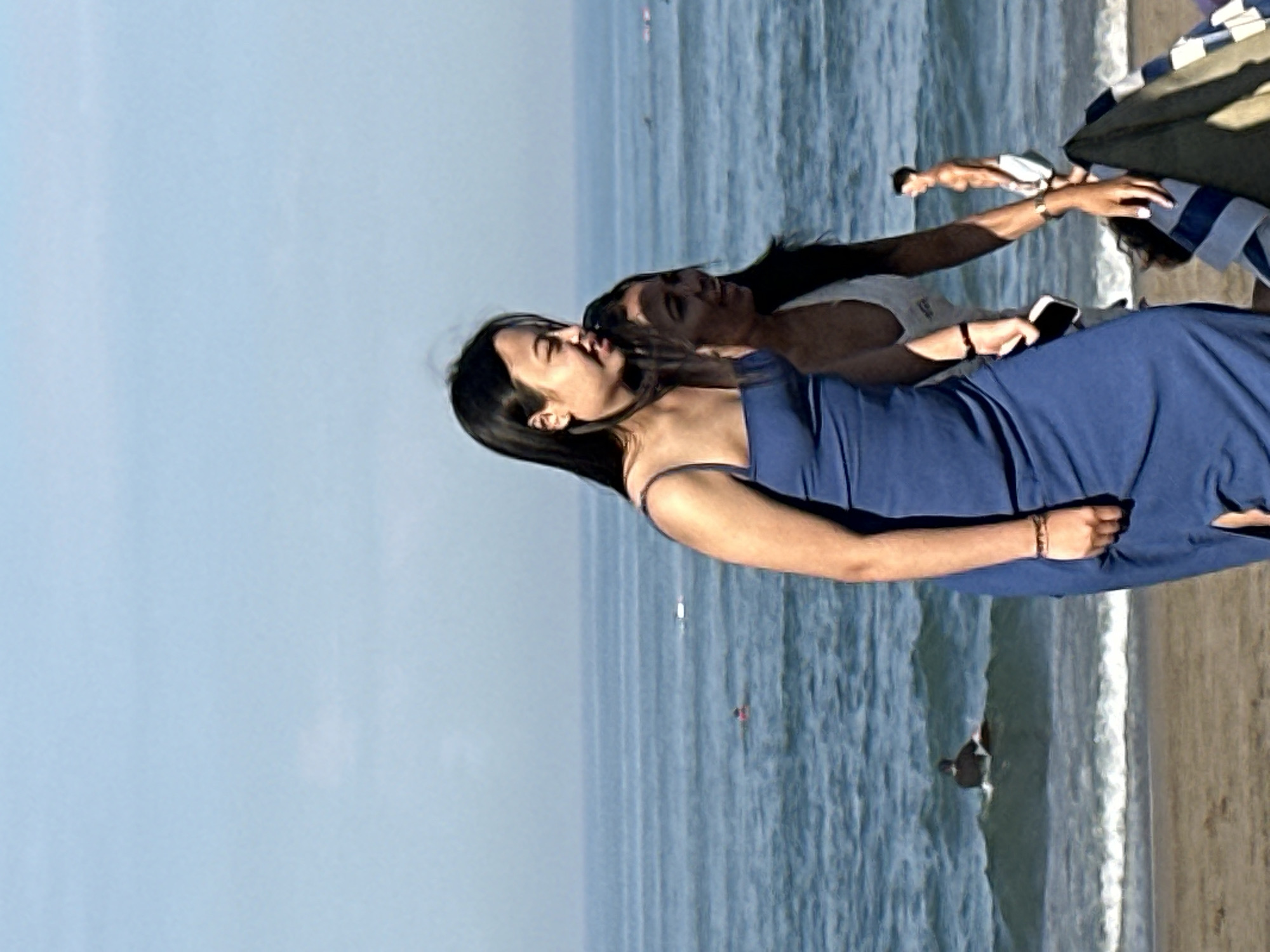 Two young women, one in a blue dress and the other in a white top, are walking on a beach. The woman in the blue dress is in the foreground, facing left and looking down. She has long, dark hair and is wearing a bracelet on her left wrist. The woman in the white top is to her right, also facing left and looking forward. The ocean is in the background, with gentle waves. A surfer is visible in the water to the left, and another person is standing on the beach to the right. The scene is set on a sunny day with a clear blue sky. The location is the beach in North Kuta, Indonesia.