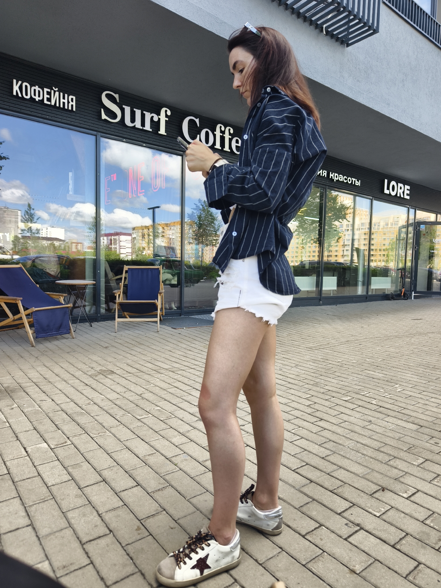 A medium shot shows a young woman with dark brown hair, wearing a navy pinstripe blazer over a white top and white distressed denim shorts, standing on a paved walkway in front of a storefront. She is looking down at a smartphone in her hands. The storefront has large glass windows and a dark facade with white lettering that reads "КОФЕЙНЯ Surf Coffe", "ия красоты LORE". Visible through the window is outdoor seating with two blue chairs and a small table. The background reveals modern buildings and a clear sky with clouds. A dark-colored electric scooter is parked near the entrance. The image appears to be taken in the city of Tyumen, Russia, based on the Russian text on the storefront.