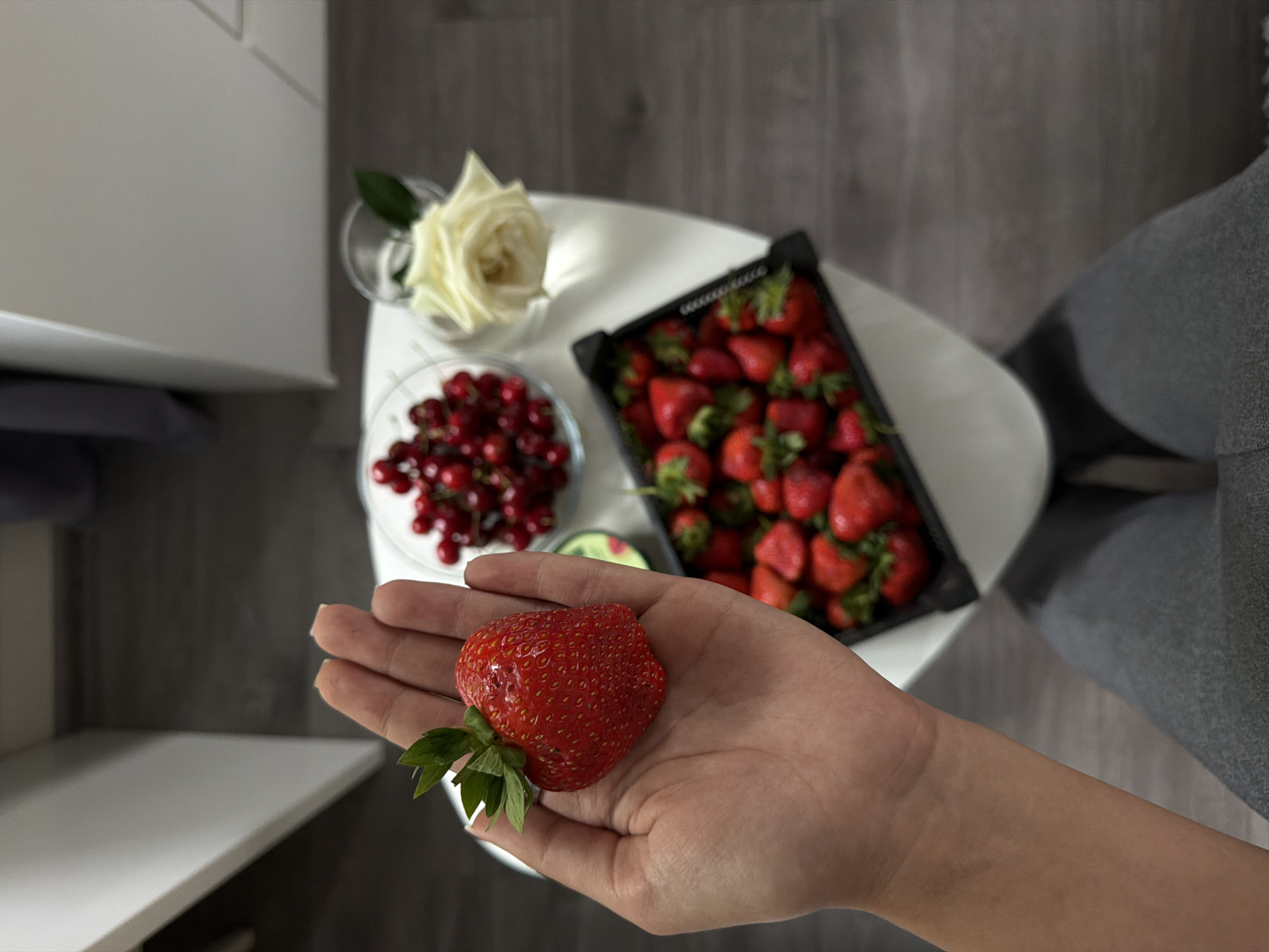 The image presents an elevated, subjective view of a scene likely indoors. A person, identifiable by their arm and hand in the foreground, is holding a large strawberry. The backdrop features a small, white, triangular table. On the table, there is a container of strawberries, a small dish filled with red currants, and a rose in a glass. Parts of white furniture and a gray chair are visible in the periphery. Based on visual clues, the location is indeterminate. However, it is explicitly stated to be Kazan, Russia.