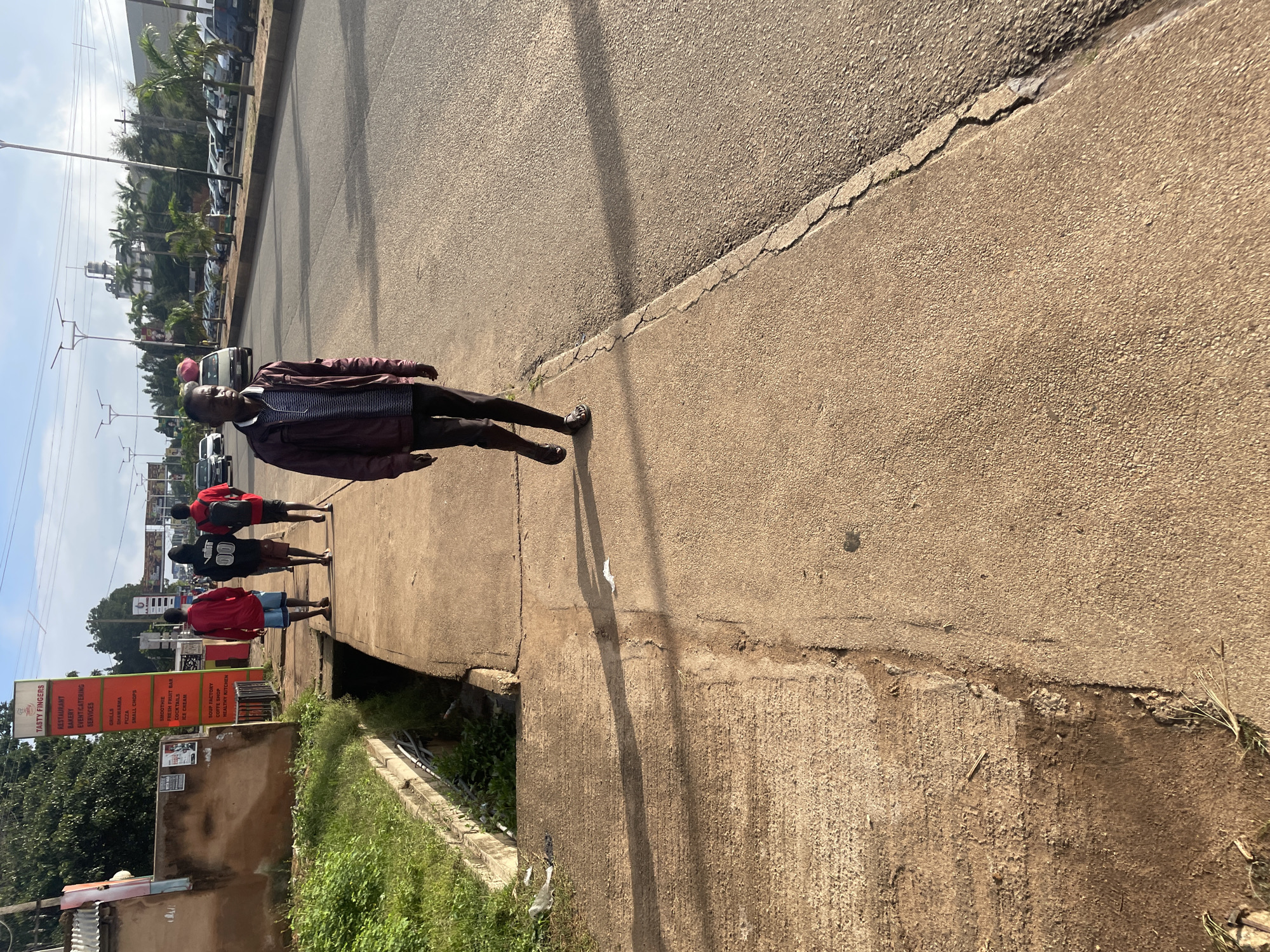 The image depicts a street scene in Jos, Nigeria, under a bright sky with scattered clouds. In the foreground, a man wearing a maroon jacket over a striped t-shirt and dark pants walks on a concrete sidewalk. He is wearing sandals. Behind him, three other individuals are walking in the same direction on the sidewalk. The first is wearing a red t-shirt and blue shorts, the second a black t-shirt with "00" on it and black shorts, and the third a red t-shirt and dark shorts. To the left of the sidewalk, there is a patch of green grass and a drainage ditch.
Further down the sidewalk, a tall red sign with white lettering, labeled "TASTY FINGERS," lists various food and service offerings such as "RESTAURANT," "BAKERY," "EVENT/CATERING SERVICES," "GRILLED SHAWARMA," "PIZZA," "SMOOTHIE," "FRESH FRUIT BAR," "COCKTAILS," "ICE CREAM," "SOUP FACTORY," "COFFEE SHOP," and "HEALTHY KITCHEN." Adjacent to the sign is a building with a brown facade.
The street itself is paved and has multiple lanes of traffic. Cars are parked along the right side of the road. In the background, buildings and palm trees are visible. The overall environment suggests a commercial area within the city.