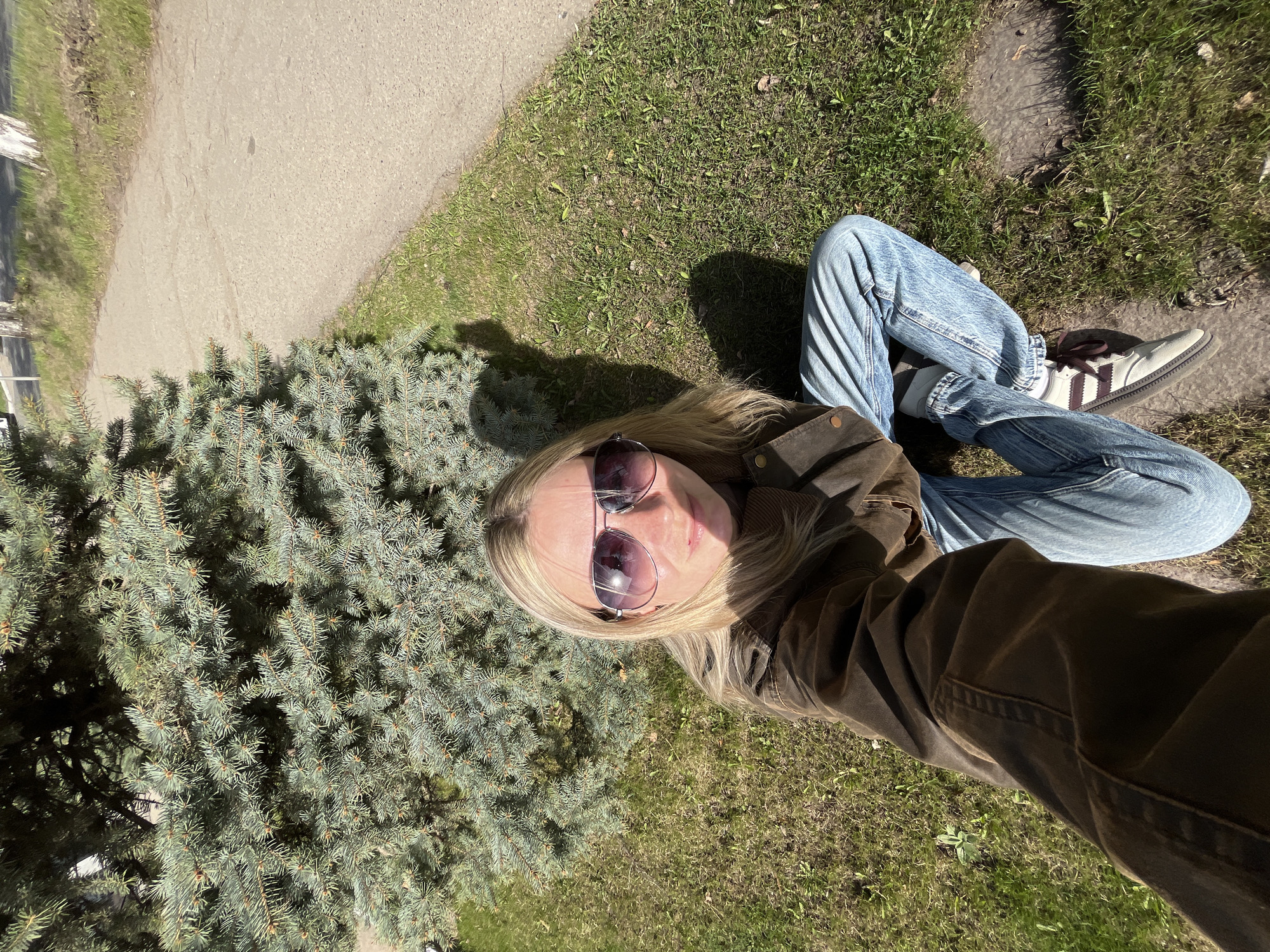 The image depicts a person sitting on the ground in an outdoor setting. They are wearing a brown jacket, light blue jeans, and sneakers. The person is also wearing sunglasses. To their left is a dense, bushy evergreen. Behind them is a grassy area, with a sidewalk visible in the upper left of the frame. The location is not possible to determine based solely on this image without additional information or context.