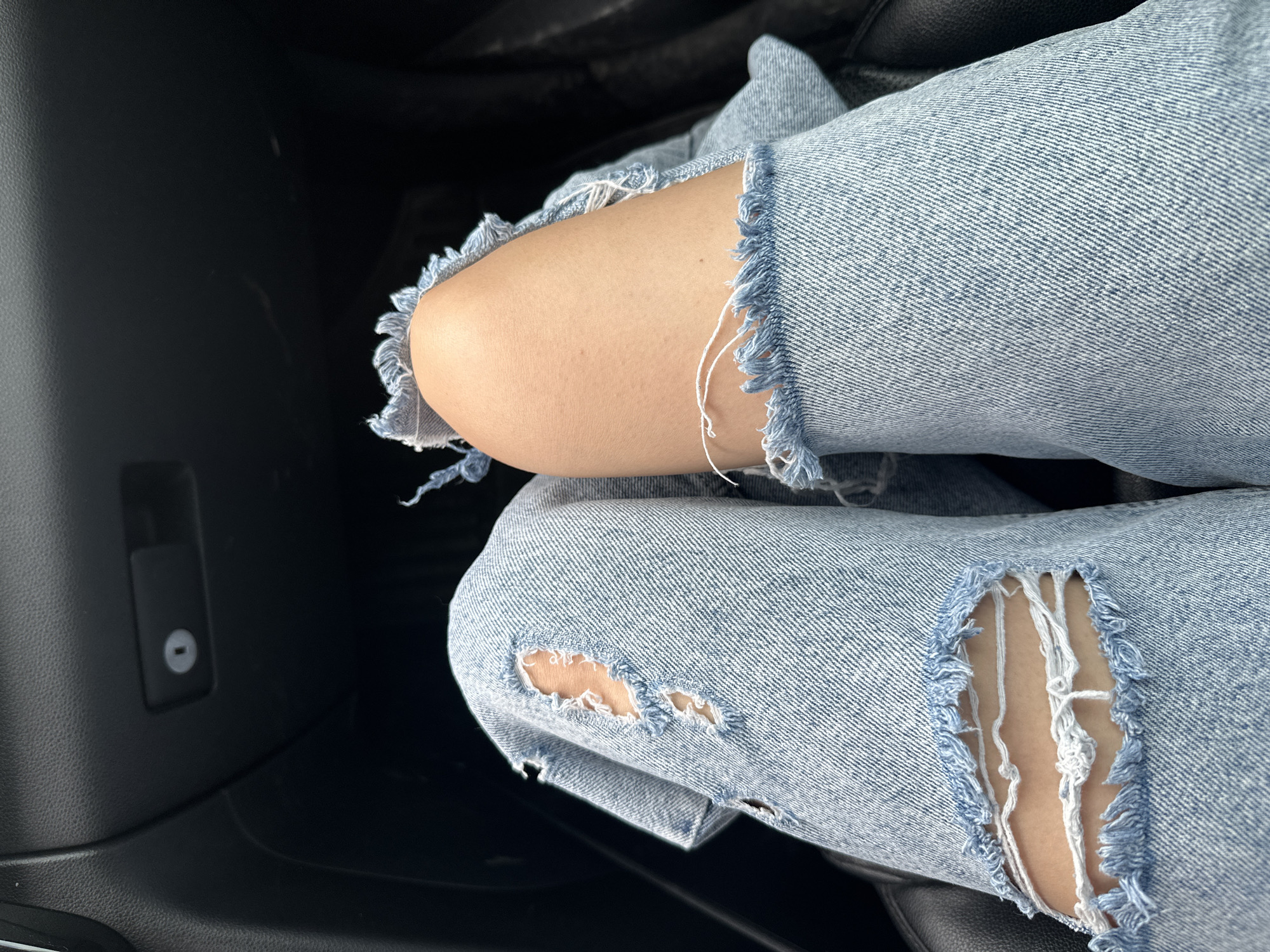 The image shows a close-up view of a person's legs wearing ripped light-wash denim jeans. The jeans have multiple tears, particularly around the knees, revealing the skin beneath. The fabric is textured with the characteristic diagonal weave of denim, and frayed threads are visible at the edges of the rips. The person appears to be sitting, possibly in a vehicle, as suggested by the dark, textured material in the background which resembles a car's interior. There is no visible indication of Yekaterinburg, Russia, or any city-specific elements within the frame.