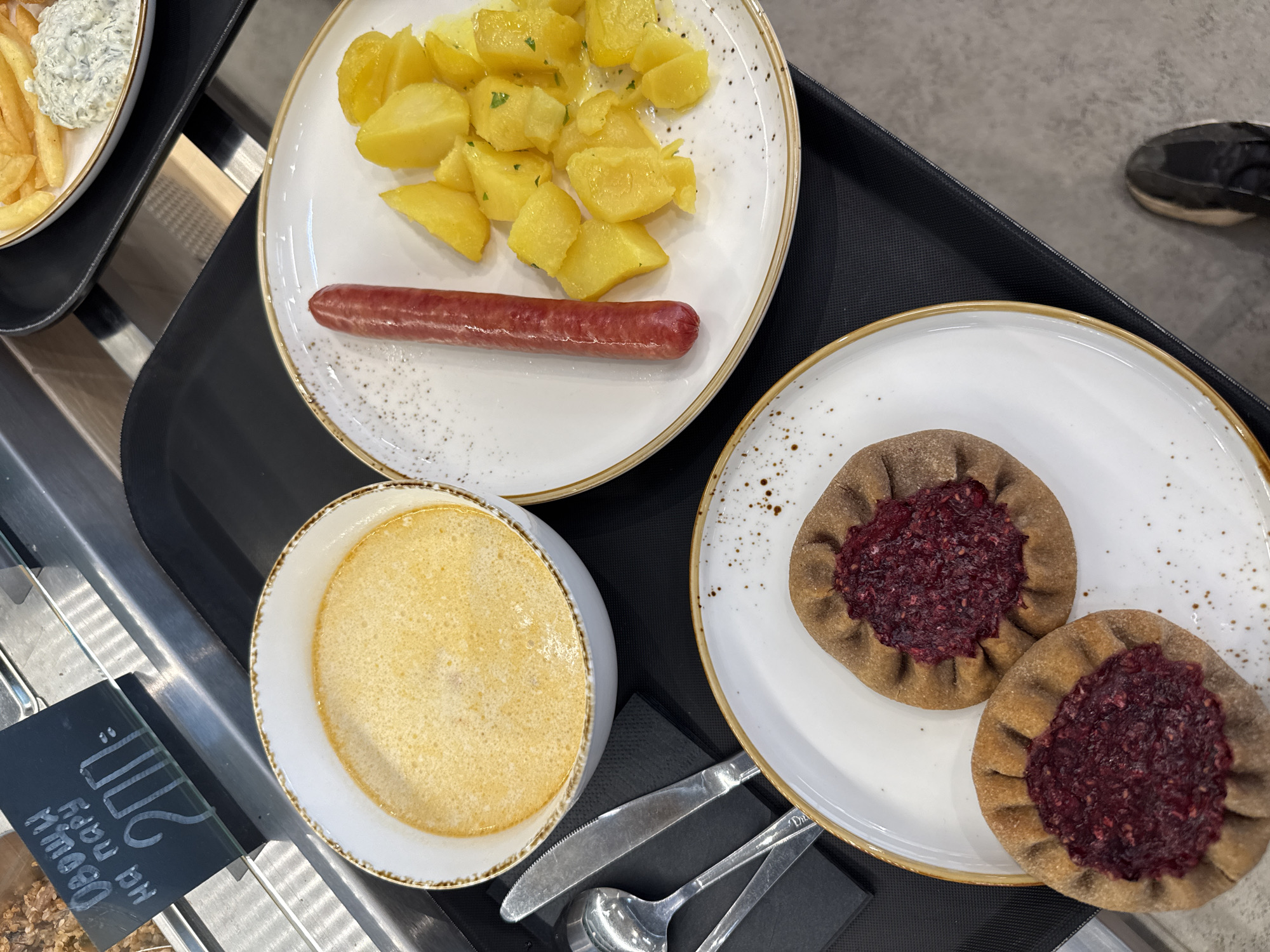An overhead, eye-level shot shows a tray of food from a cafeteria in Yakkima, Russia. The tray holds a white bowl of creamy yellow soup, a white plate with a single sausage and a serving of diced yellow potatoes, and a white plate with two small, round pastries filled with a dark red berry mixture. A fork and knife are laid out on a black napkin next to the soup. A black sign above the soup reads "Овощи на пару 200" which translates to "Vegetables for two 200". A pair of dark sneakers is visible in the lower right corner of the frame. The background appears to be a stainless steel display case.