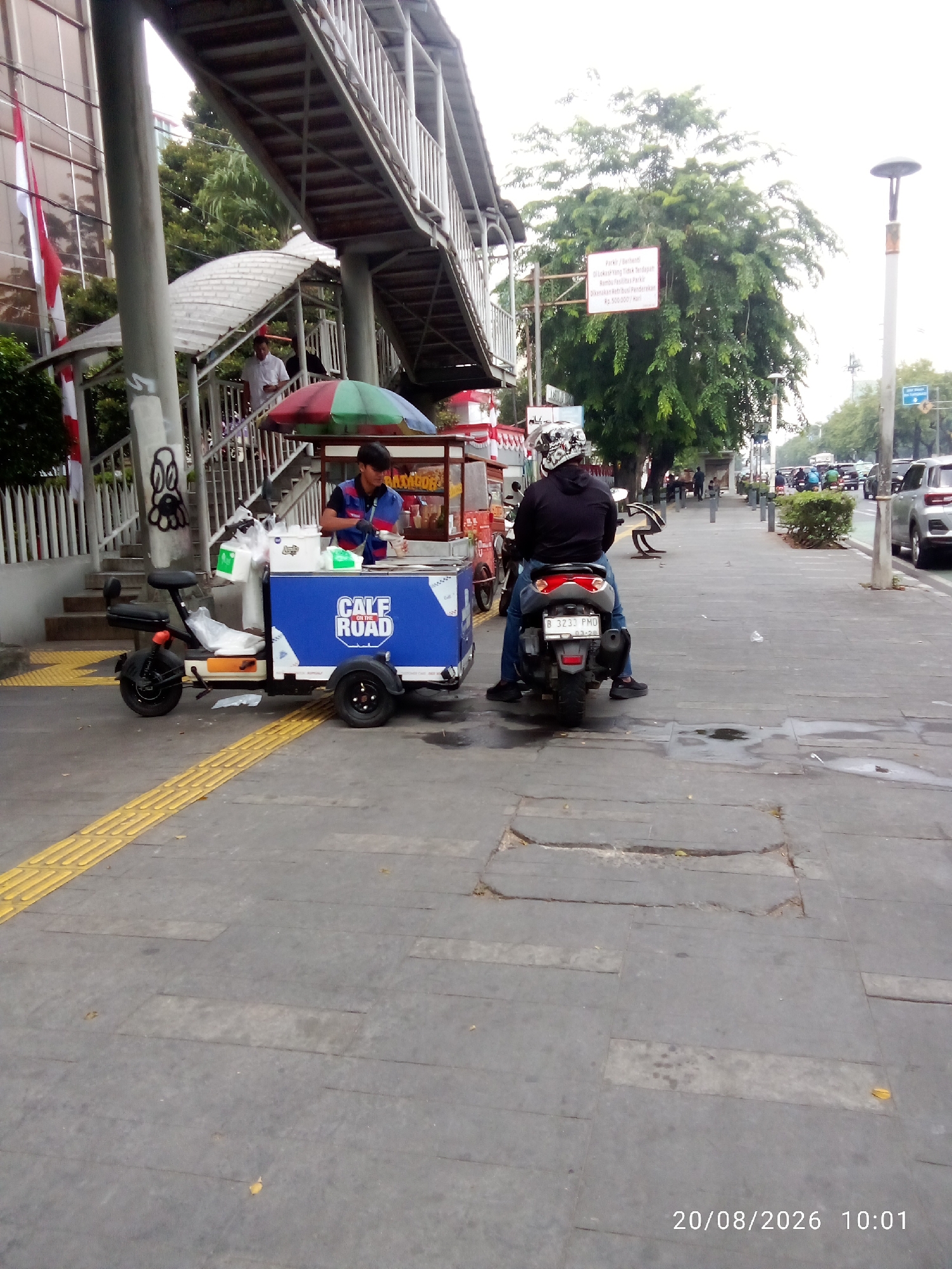 In Central Jakarta, Indonesia, an outdoor scene unfolds with a street vendor's cart as the central focus. The cart, painted blue with white lettering that reads "CALF ON THE ROAD", is equipped with a green and red umbrella, suggesting it's an ice cream or beverage stand. A man, presumed to be the vendor, is actively serving a customer from behind the cart, handling what appears to be a drink or food item.
In front of the cart is a small electric scooter, also blue and white, parked and ready for transport. To the right of the vendor's cart, a person is seated on a motorcycle, facing away from the camera, indicating they are either waiting or have just arrived. The motorcycle has a license plate that reads "B 3233 PMD".
The background reveals elements typical of an urban environment. A pedestrian overpass with metal railings and concrete supports runs overhead, hinting at a busy intersection or thoroughfare. The overpass structure is partially obscured by lush green trees. A building with vertical windows is visible on the left side, adorned with Indonesian flags. The ground is paved with concrete tiles, featuring a yellow tactile paving strip along the edge of the walkway. The weather appears to be overcast, as indicated by the diffused light and the overall bright yet soft illumination of the scene. The timestamp "20/08/2026 10:01" is overlaid on the bottom right of the image.