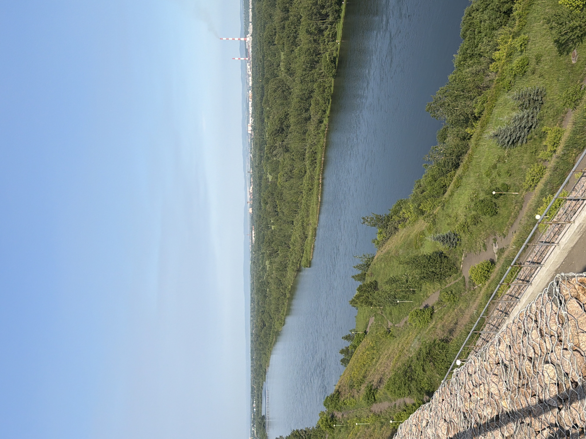 This is a landscape photograph taken from a high vantage point, overlooking a wide river and a densely forested embankment. The sky is a clear, pale blue, suggesting a bright, sunny day. In the distance, a city skyline is visible, with several industrial chimneys emitting plumes of white smoke, indicating industrial activity. The river is a deep blue and appears calm, reflecting the sky. The embankment on the right side of the river is covered in lush green vegetation, with trees and bushes growing on a sloping hillside. A paved path winds through the greenery, and a metal railing runs along the edge of the viewpoint. In the foreground, a gabion wall made of large stones enclosed in wire mesh is visible, suggesting the location is a developed area, possibly a park or viewing platform. The scene conveys a sense of natural beauty combined with urban development. No people or animals are visible in the image. The time of day appears to be daytime, given the clear sky and sunlight. The location is identified as Krasnoyarsk, Russia, which is consistent with the presence of a large river and industrial infrastructure.