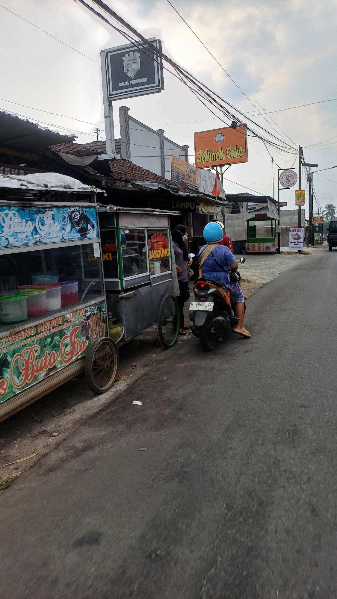 The image depicts a street scene in Tengahan, Indonesia, with several businesses and a food cart visible. In the foreground, a food cart labeled "Buto Ijo" is parked on the side of the road. The cart has colorful graphics and displays containers of what appear to be drinks. To the right of the food cart, two individuals are interacting. One person, wearing a blue helmet, is seated on a motorcycle. The motorcycle has a license plate reading "H 4069 ZC". Another individual, wearing a headscarf and mask, stands near the food cart.
Further down the street, a prominent orange sign displays "Sakinah Cake". Above this, a black and white sign reads "BAJA PERFUME". There are various other shopfronts and signs in the background, some displaying text in Indonesian. The road surface is asphalt, and the scene is under an overcast sky. The overall impression is of a bustling local commercial area.