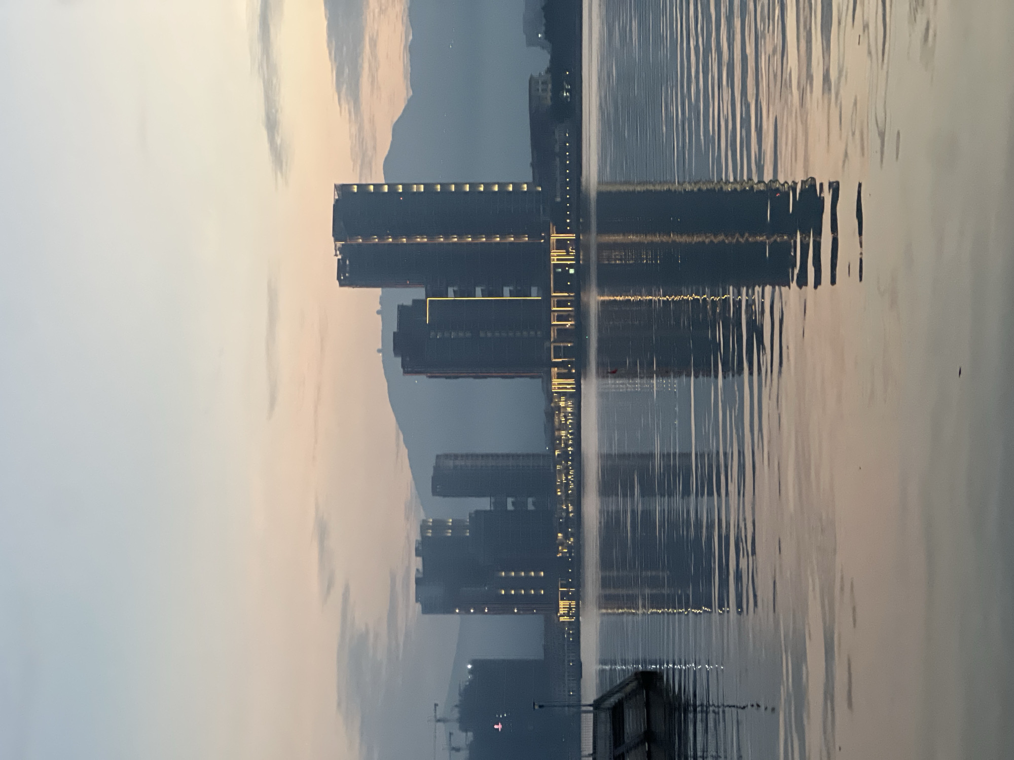 The image captures a serene morning view of the Dragon Bridge area in Da Nang, Vietnam. In the foreground, calm water reflects the soft hues of the dawn sky, which transitions from a pale blue at the top to a warm pink and orange near the horizon. Several modern, multi-story buildings with illuminated windows stand on the far bank, their reflections shimmering on the water's surface. One prominent cylindrical building is particularly striking. A construction crane is visible to the left of the buildings, indicating ongoing development. A wooden pier with steps leads into the water on the left side of the frame. The background features misty mountains that are partially obscured by the atmospheric haze. The scene is tranquil, devoid of people, and evokes a sense of early morning quietude in a developing urban landscape.