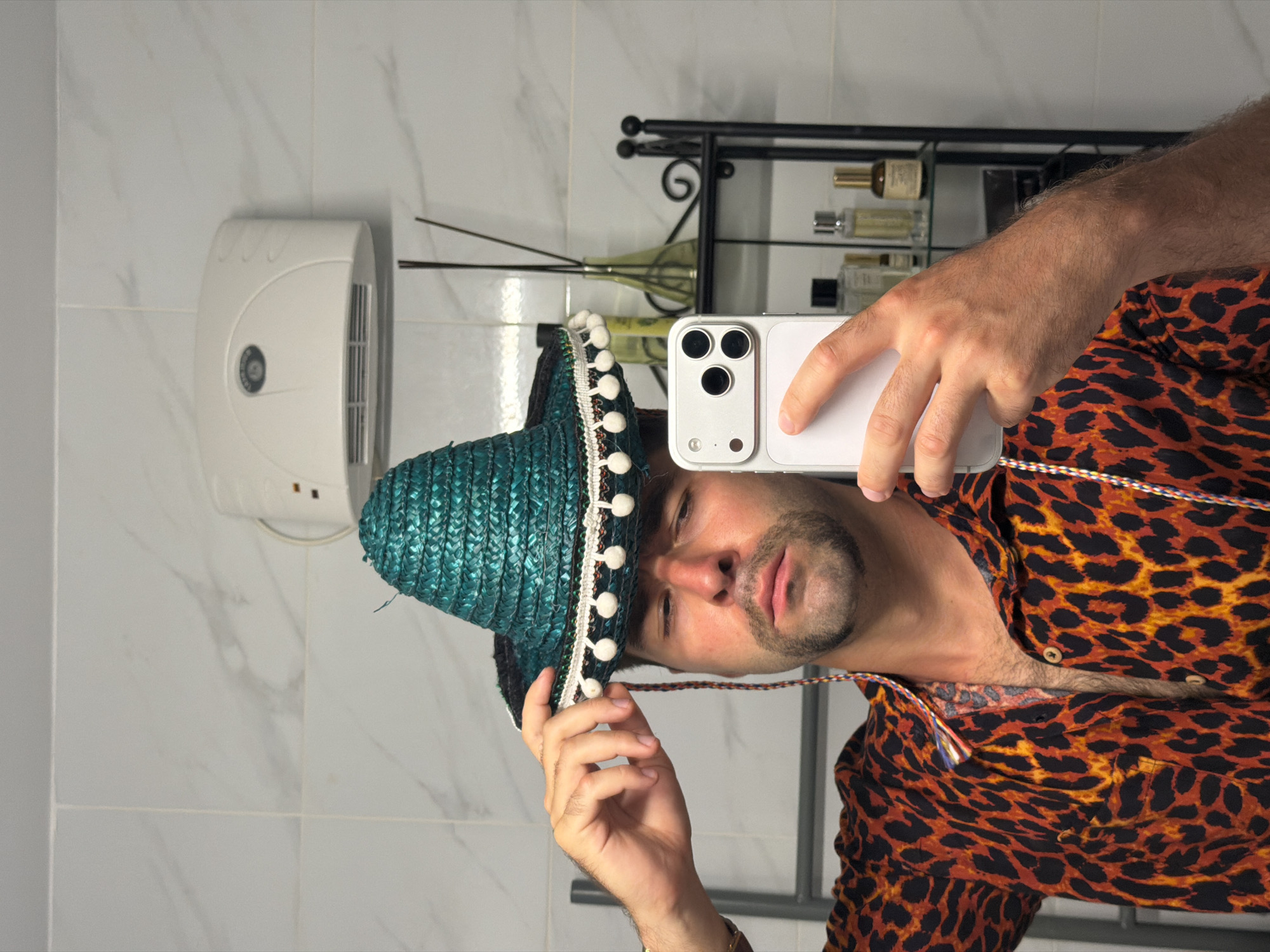 A man is taking a selfie in a bathroom. He is wearing a teal sombrero with white pom-poms and a colorful patterned shirt. In the background, there is a shelf with bottles and a towel rack. The bathroom appears to be in an apartment in Tel-Aviv, Israel, based on the tiled walls and general aesthetic.