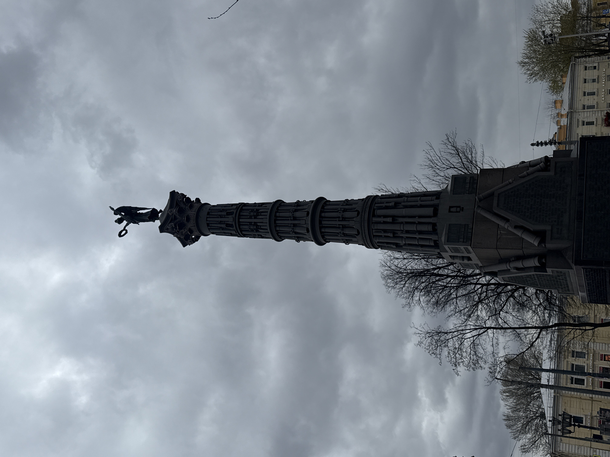 The image captures a tall, dark-colored monumental column under an overcast, gray sky. The column, viewed from below, is intricately designed, appearing to be composed of stacked cylindrical elements that resemble bundled fasces or cannon barrels. Perched atop the column is a statue of a winged figure, likely an angel or goddess of victory, holding a wreath or similar object, silhouetted against the bright sky.
Bare-branched trees surround the monument, suggesting a late winter or early spring season. In the urban background, several buildings are visible, including a multi-story light-colored structure with numerous windows. Overhead electrical wires or tram lines crisscross the scene, and a traffic light pole can also be seen.
Cyrillic text is discernible on one of the background buildings: "САЛОН КРАСОТЫ" (Beauty Salon) and "КОФЕ ИЛИ СТРИТФУД" (Coffee or Street Food), along with "РИЧЕЛЛО". These textual and architectural cues, combined with the style of the monument, firmly place the scene in Saint Petersburg, Russia. The cloudy conditions indicate a dull daytime, without direct sunlight. There are no people visible, and no specific event is taking place, emphasizing the monumental and historical presence of the column.
