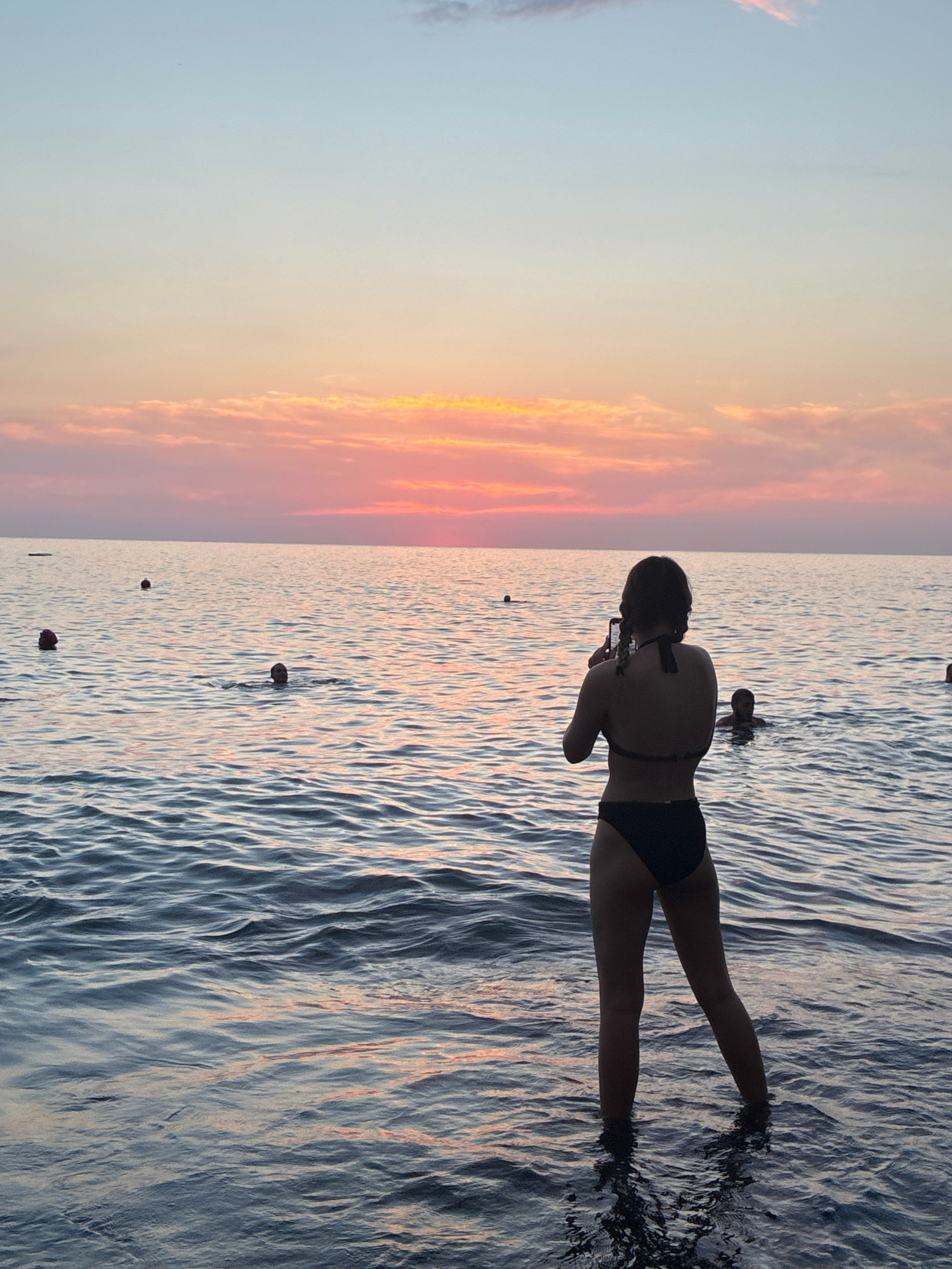 A young woman wearing a black bikini stands in shallow water at sunset, holding a smartphone to take a photograph. The water is calm with gentle ripples, reflecting the soft colors of the sky. In the background, several people are visible in the water, some swimming and others treading water. The sky transitions from pale blue at the top to hues of pink, orange, and yellow near the horizon, indicating a sunset. The overall scene suggests a peaceful evening at a beach, likely on the coast of Sochi, Russia, given the context.