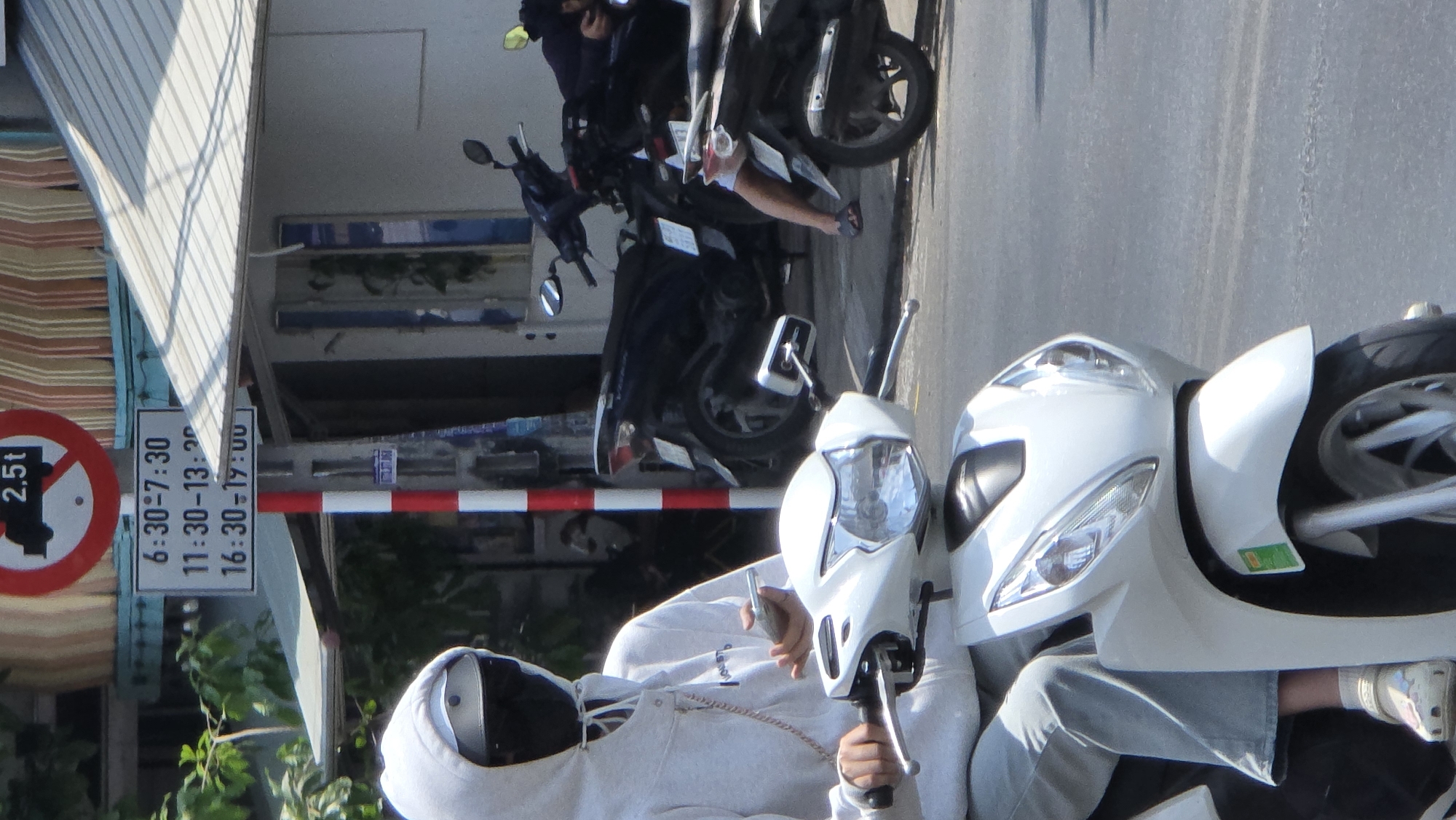 The image depicts a street scene in what appears to be a city, possibly Bắc Nha Trang, Vietnam. The focal point is a person on a white scooter. The person is wearing a grey hoodie, jeans, and a helmet, with the hoodie pulled up over their head. The person seems to be holding the handlebars and seated on the scooter.
In the background are more scooters, some black and others with unidentified colors. There is a sign indicating weight restrictions ("2,5t") with time restrictions written below. A red and white striped barrier is present, and urban structures can be seen, including what might be a building facade. The street seems to be paved, and daylight is present.