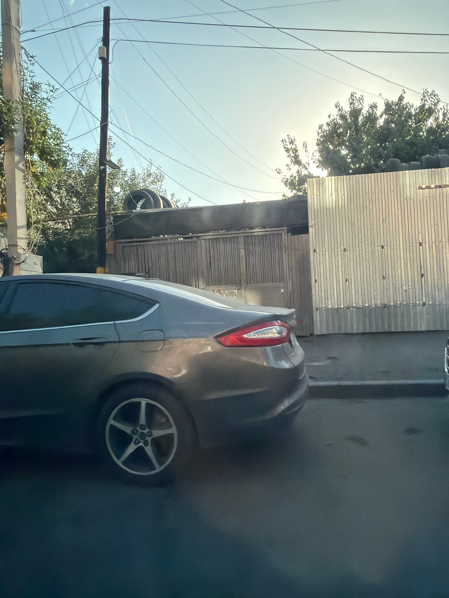The image depicts a street scene with a grey sedan parked on the right side of the road. The car is positioned at an angle, facing away from the camera, with its rear visible. The vehicle appears to be a modern sedan with tinted windows and alloy wheels. In the background, there is a fence constructed from corrugated metal panels, and to the left, a wooden gate with vertical slats. Overhead, a power pole with numerous wires stretches across the frame, indicating an urban environment. Trees with green foliage are visible behind the fence and gate. The sky is clear and bright, suggesting daylight. There are no people visible in the frame. The scene appears to be from a residential or industrial area within Yerevan, Armenia, characterized by the presence of utility poles and fencing typical of such locations.