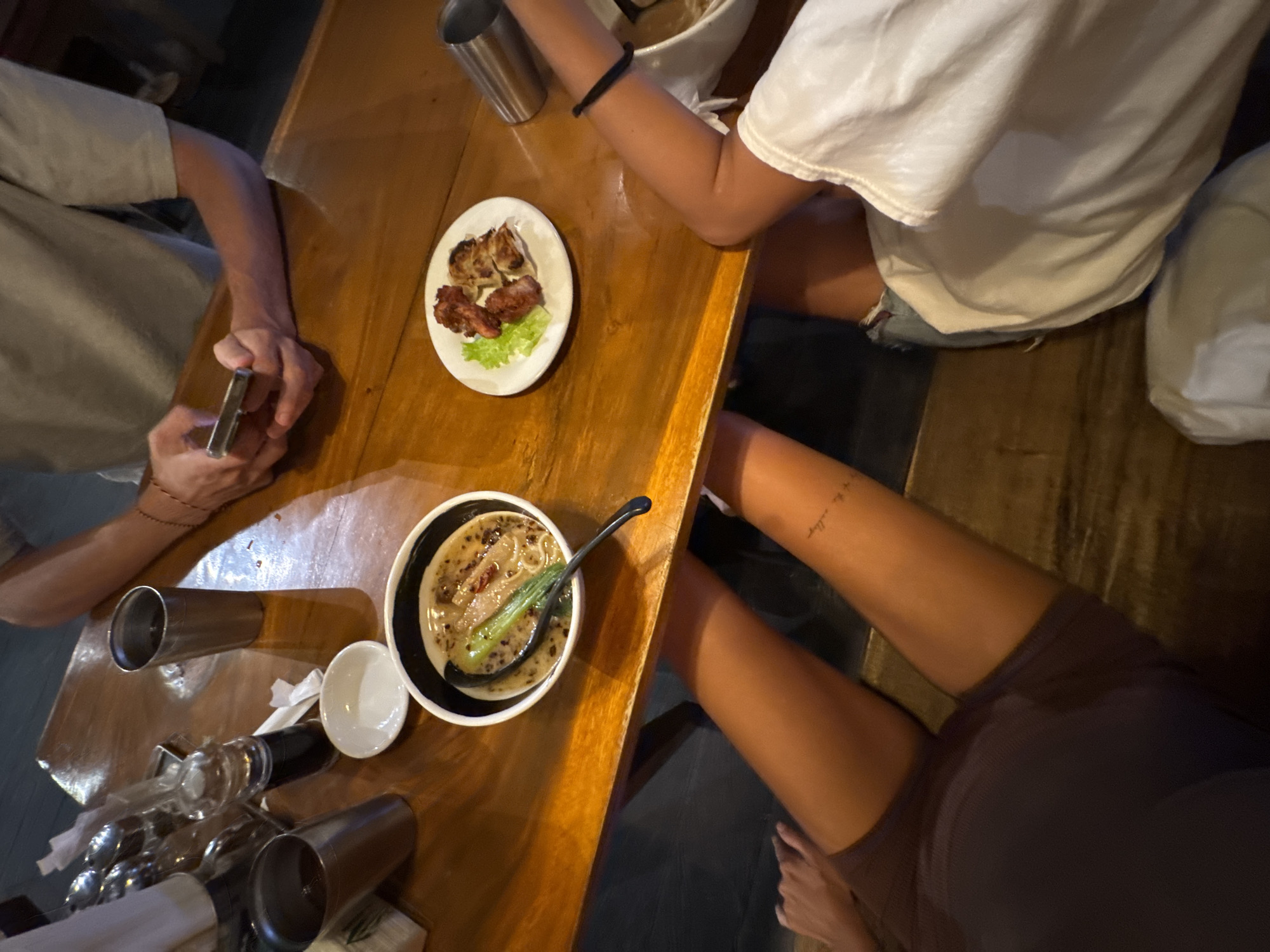 Here is a description of the image:
The image captures a dining scene in what appears to be a restaurant. The focal point is a wooden table on which various food items and dining accessories are placed. A person in a white t-shirt sits to the right of the table, while another person with a grey shirt on the left is using a smartphone.
On the table, there's a plate with a few pieces of meat and some greens. A bowl of soup or noodles with a spoon resting inside can also be seen. Other objects on the table include glasses, a bottle, and a small dish.
One person wears a black band on their arm. Another person has tan legs and is wearing a dark purple dress. The location is North Kuta, Indonesia.