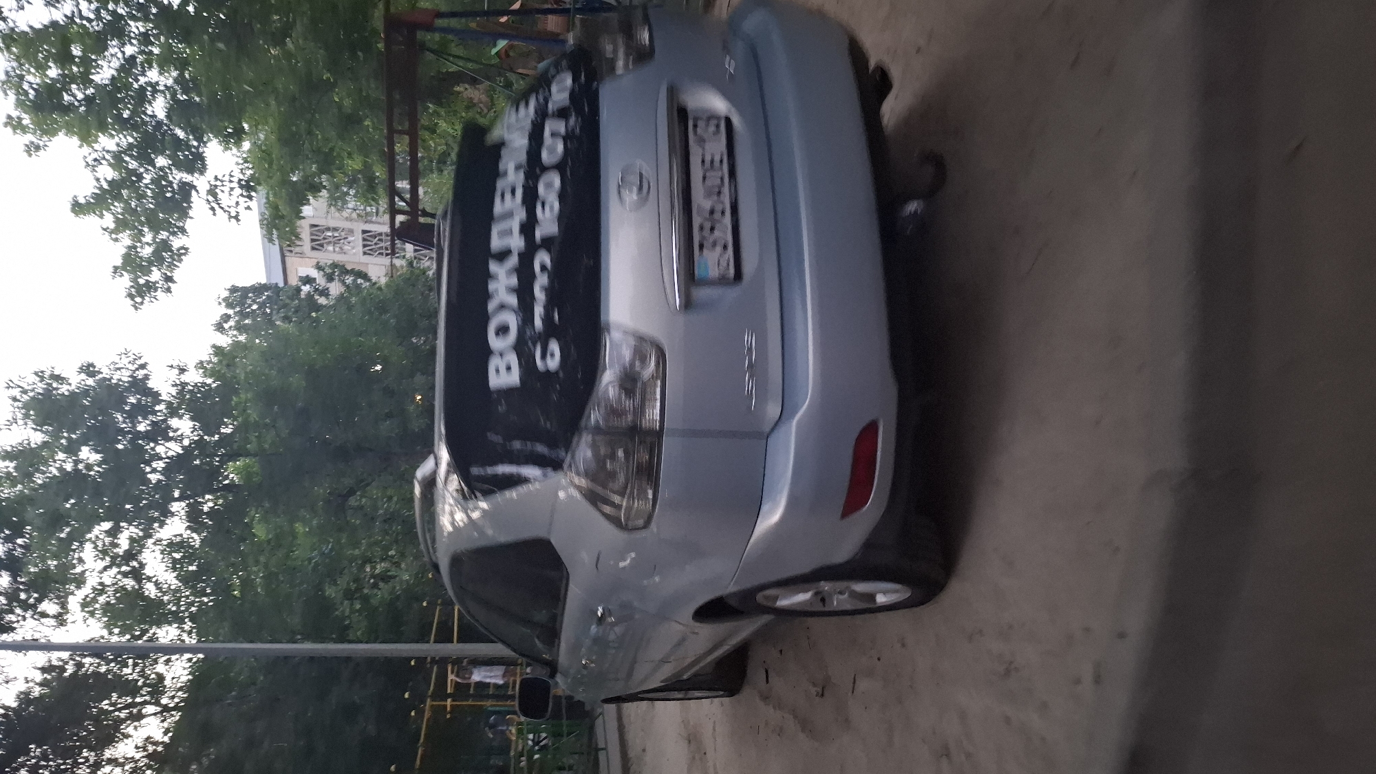 A silver Lexus RX is parked on a dirt surface in front of a building and trees. The vehicle has a black decal on the rear window with white lettering that reads "ВОЖДЕНИЕ" and "6 702 160 07 10". The license plate is visible and appears to be a Kazakh plate with the characters "396ADE13". There is a swing set visible in the background to the right of the car. The overall scene appears to be in a residential area of Shymkent, Kazakhstan, possibly a courtyard or parking lot.
