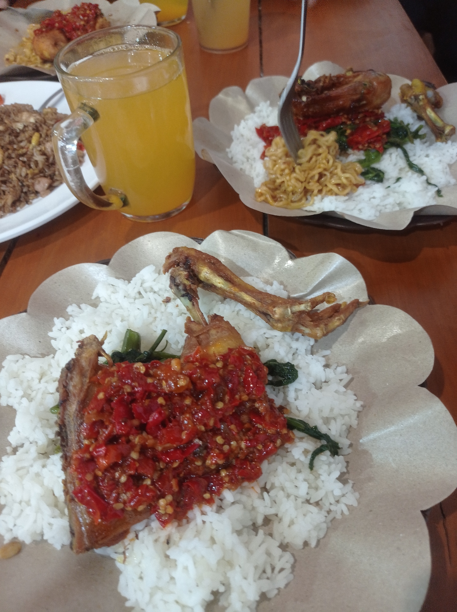 The image depicts a close-up, overhead view of a wooden table in a restaurant, likely in Landungsari, Indonesia. On the table are several plates of food and a glass of orange juice.
In the foreground, a plate of white rice is topped with a piece of fried chicken covered in a red chili sauce. Some green vegetables are mixed in with the rice. To the left of this plate, another plate contains fried rice and a dish with red toppings.
In the middle ground, another plate of white rice is served with fried chicken, a small portion of fried noodles, and red chili sauce. A fork is visible, partially inserted into the noodles. A glass of orange juice, with ice, is situated to the left of this plate.
In the background, a blurry orange beverage is visible in a glass. The lighting suggests an indoor setting. The overall impression is of a casual meal at a restaurant.
