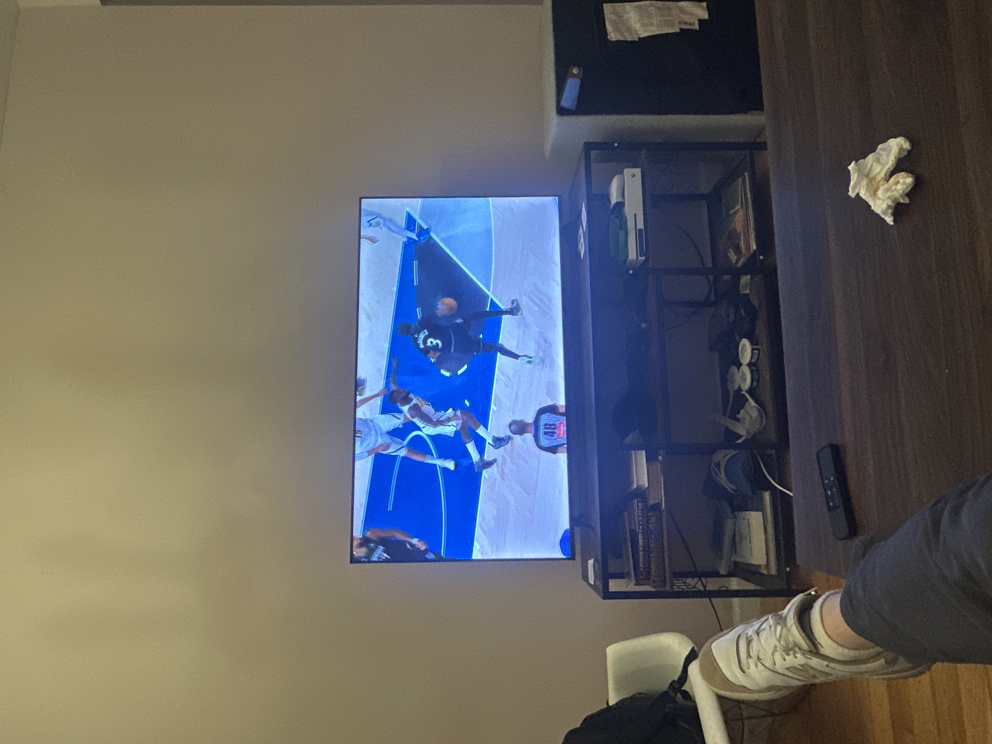 An indoor scene shows a flat-screen television displaying a basketball game, mounted on a light-colored wall. The image is oriented vertically, showing the TV on the left, a media console and wooden surface on the right, and a person's foot in the bottom right foreground.
On the television, several basketball players are visible on a court with blue markings. One player in a dark jersey with the number "3" is dribbling the ball, while another player in a white jersey and a player in a dark jersey with "48" are also on screen. The game appears to be in progress.
To the right of the television is a black metal media console or shelving unit. On one shelf, a white Xbox console is clearly visible. Below it, several books and various cables are stored. To the right of the console, on a dark wooden surface, a black remote control is placed, and a crumpled white tissue or piece of paper is also visible.
In the bottom right corner, a person's leg and foot are partially visible, wearing light-colored New Balance sneakers with tan accents and dark pants, suggesting a relaxed posture. A white chair is partially obscured next to the foot. The room features plain light-colored walls and a dark wooden surface that could be a desk or floor. The lighting suggests an indoor setting, possibly evening or night, with artificial light sources. No specific location cues for New York are visible within the image itself.