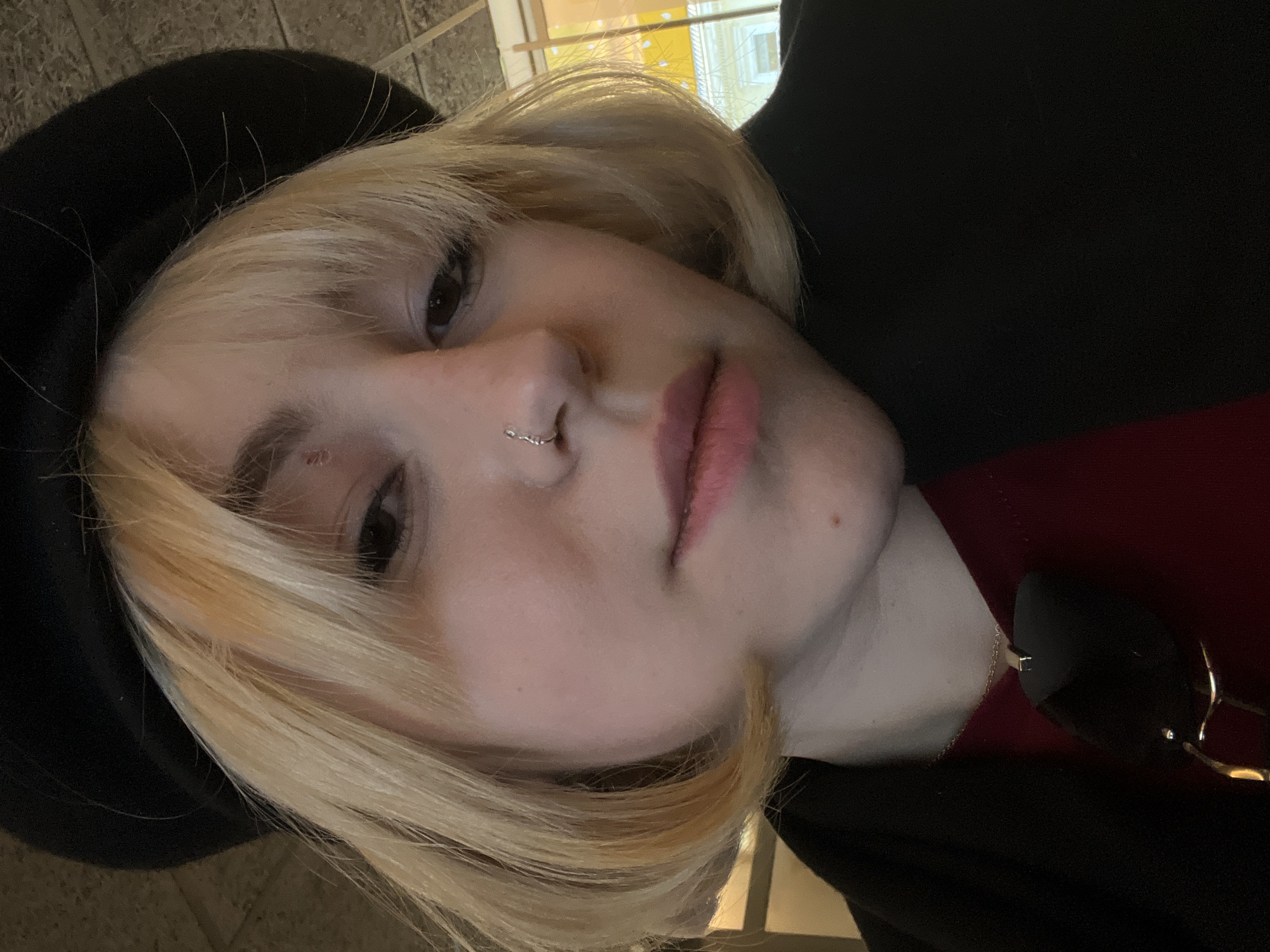 A close-up, rotated selfie features a young person with light blonde, bob-length hair and fair skin. The individual has dark eyes and is looking directly at the camera with a neutral to slightly pensive expression. A small silver ring piercing adorns their nose. They wear a dark, possibly black, beret or hat, which covers the top of their head. Visible attire includes a red garment, likely a shirt or sweater, layered under a dark, possibly black, outer jacket or coat. A pair of sunglasses with dark lenses and a thin gold-colored frame is seen resting on the red garment.
The background is largely dark and out of focus, suggesting either an indoor setting or an outdoor environment at night or dusk. Some indistinct light sources or reflections are visible in the upper background. No specific location cues are present within the image, though the provided context indicates the location is Odesa, Ukraine. The overall lighting appears dim, contributing to a subdued atmosphere. There is no visible text in the image.