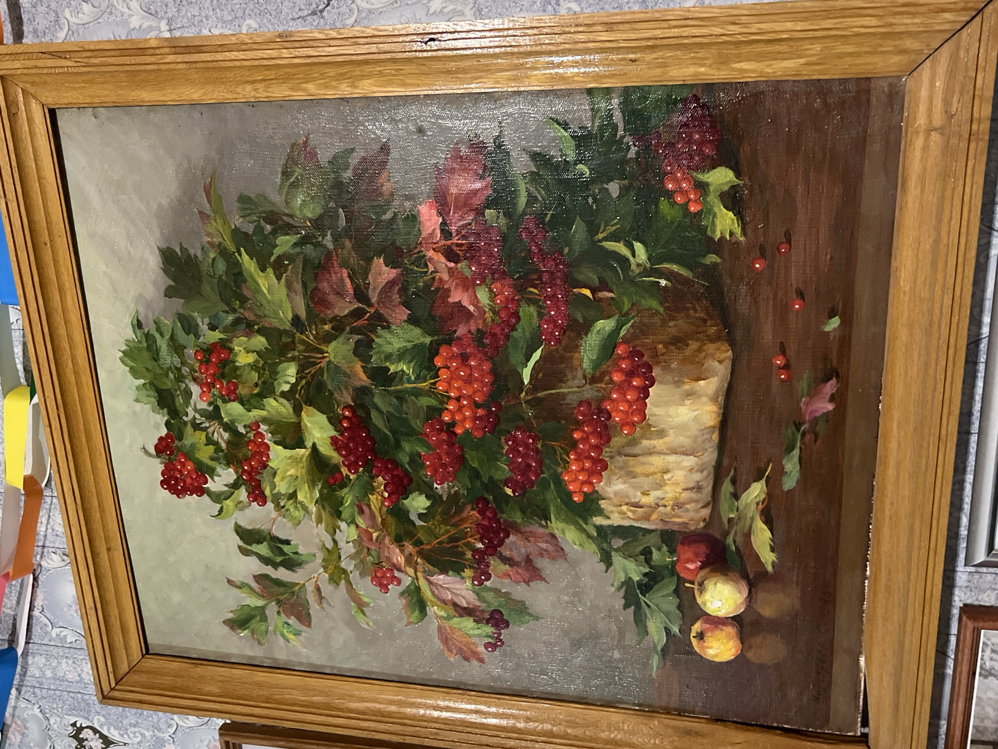 This image features a still life oil painting enclosed in a wooden frame. The painting depicts a vibrant arrangement of red berries, possibly viburnum or rowan, presented in large clusters alongside green and some autumnal reddish-brown leaves. The berries and foliage are gathered in what appears to be a light-colored, possibly woven or bark-textured, basket or vase. The background transitions from a soft, muted grey-green on the left to a dark brown surface, resembling a wooden tabletop, on the right.
On the dark brown tabletop, several individual red berries are scattered, and near the bottom right, two small apples are visible – one predominantly red and the other yellowish-green with red blush. The painting exhibits a realistic style with visible texture, indicative of oil on canvas. The frame is made of light-colored wood, likely oak, featuring a simple, grooved profile. In the bottom right corner of the painting, a signature can be discerned, reading "А. Васюков 88," suggesting it was created by A. Vasyukov in 1988. The overall image captures the painting resting on a light blue-grey patterned surface. The context for the image is Новая Тура, Russia.