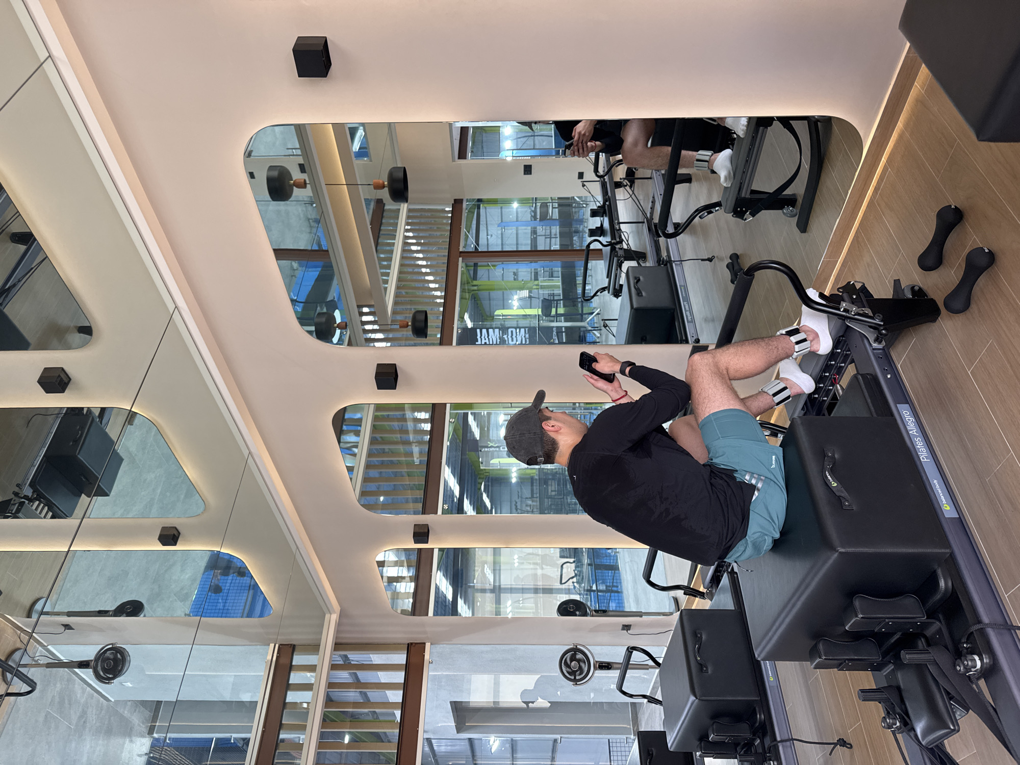 The image captures a modern Pilates studio, likely located in San Bernardino Tlaxcalancingo, Mexico, from a high-angle, side perspective. The primary subject is a man seated on a Pilates reformer machine. He wears a black long-sleeved shirt, teal athletic shorts, a dark baseball cap, and white socks with ankle weights. He is actively engaged with his cell phone, holding it in both hands and looking down at the screen, suggesting a break, checking instructions, or recording his workout. A slight sheen of sweat is visible on his neck, indicating recent physical exertion.
The Pilates reformer itself is dark grey or black, with the brand "Pilates Allegro" and "Balanced Body" visible on its side. Several small, black, ergonomic dumbbells are placed on the light wood-patterned floor next to the machine.
The studio's interior design is contemporary, featuring light-colored walls and ceiling with distinctive, organically shaped cutouts that house large mirrors. These mirrors reflect various parts of the gym, including another person (likely the same man or an instructor) on a reformer, large windows that allow natural light into the space, and bright interior lighting. Through the reflections, elements like window blinds, a floor fan, and the general open layout of the gym are discernible. Small, black, cube-shaped lights are mounted on the walls or ceiling. In one mirror reflection, a reversed sign that appears to read "JAMOW!" is visible. The overall ambiance suggests a clean, well-lit, and professionally equipped fitness environment.