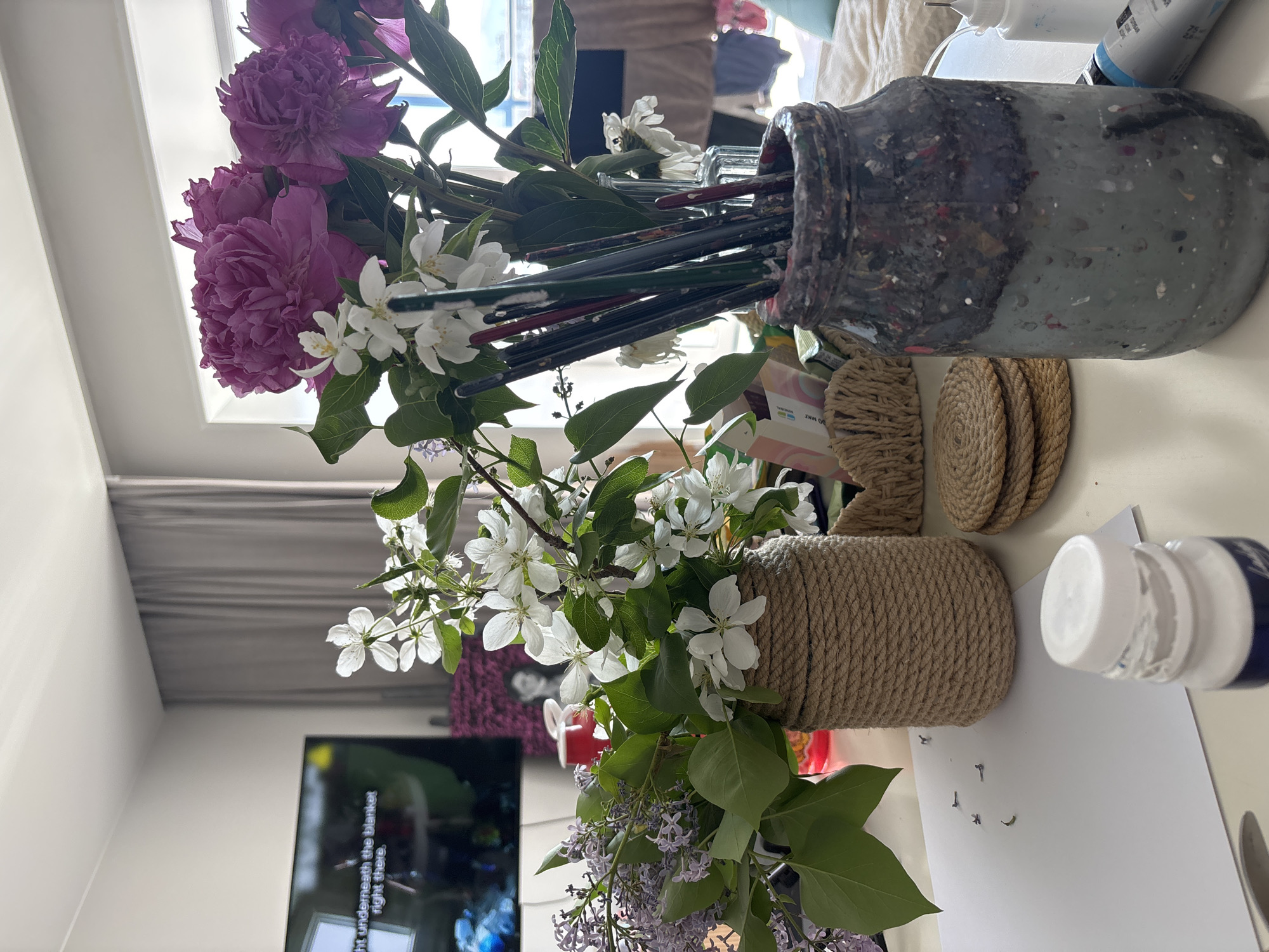 This media file captures a brightly lit indoor scene, likely a domestic setting such as a home in Krasnoyarsk, Russia. The image is oriented vertically and displays a collection of fresh flowers and art supplies arranged on a light-colored surface, possibly a table.
In the upper left, vibrant purple peonies dominate, accompanied by branches of white blossoms, likely apple or cherry. To the right, a large glass jar, heavily stained with various paint colors, holds numerous paintbrushes. Behind this jar, several tubes or bottles of paint, one showing "75 m" (possibly milliliters), are visible. Adjacent to the paint jar are neatly stacked round coasters made of coiled jute rope, and a larger cylindrical container also wrapped in jute, which holds more white blossoms and sprigs of light purple lilac.
In the lower right, a white bottle, possibly containing paint or glue, rests near a white sheet of paper on which a small pile of detached lilac florets is scattered. The background reveals a bright window or skylight, indicating natural daylight. In the lower left, a television screen is partially visible, displaying text that reads "...ght underneath the blanket right there." Various pieces of furniture or textiles are subtly blurred in the background, reinforcing the home environment.
The scene suggests a relaxed, perhaps creative, atmosphere, bathed in natural light. No people are visible, and no specific activity is clearly underway, but the arrangement implies a moment of stillness or preparation for an artistic endeavor.