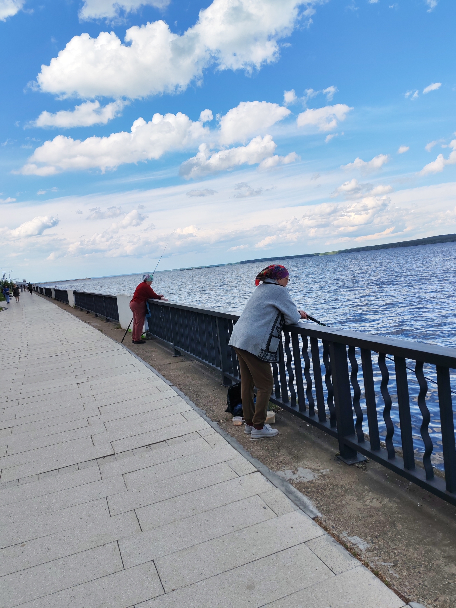 In Chaikovsky, Russia, two individuals are seen fishing from a waterfront promenade on a bright, cloudy day. The scene depicts a wide river or large body of water to the right, with a distant, tree-lined shore visible on the horizon. A paved promenade, made of light-colored rectangular tiles, stretches along the left side of the image. A black metal railing separates the promenade from the water.
The subject in the foreground is an older person, viewed from behind, wearing brown pants, grey jacket, and a colorful headscarf. They are leaning on the railing and holding a fishing rod, looking out at the water. Another person, dressed in red pants and a red top, is further down the promenade, also fishing with a rod.
The sky is a vibrant blue with scattered white cumulus clouds, suggesting fair weather and possibly late morning or early afternoon. The overall mood is peaceful and leisurely, with the individuals engaged in the quiet activity of fishing. No visible text is present in the image.