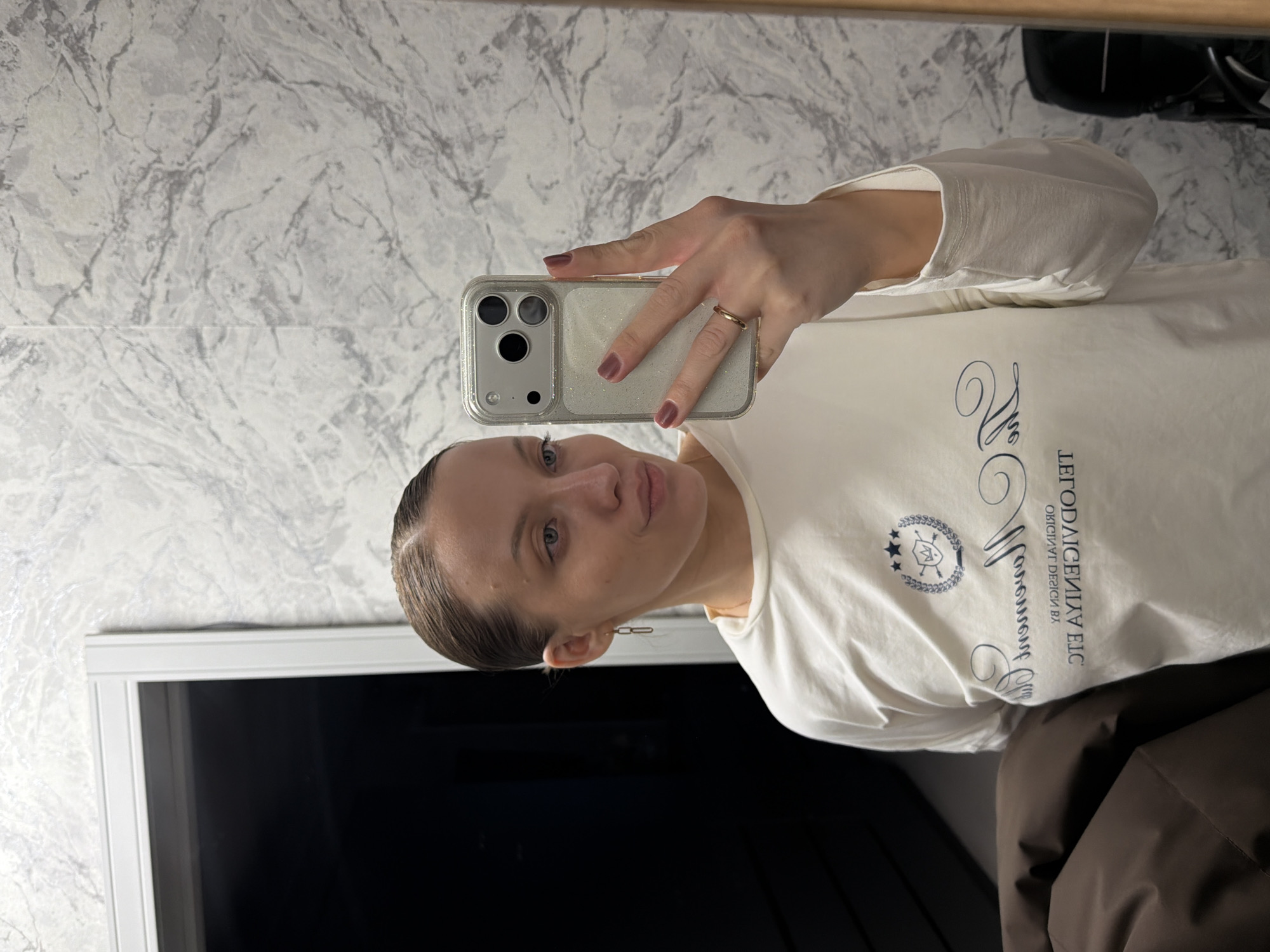 A woman is taking a selfie in front of a window. The background consists of a marble-patterned wall. The woman is wearing a white long-sleeved shirt with a logo on it. She is holding a smartphone with a glittery case. The setting appears to be an interior space. There is no indication of Reshetnikovo, Russia, in the image.