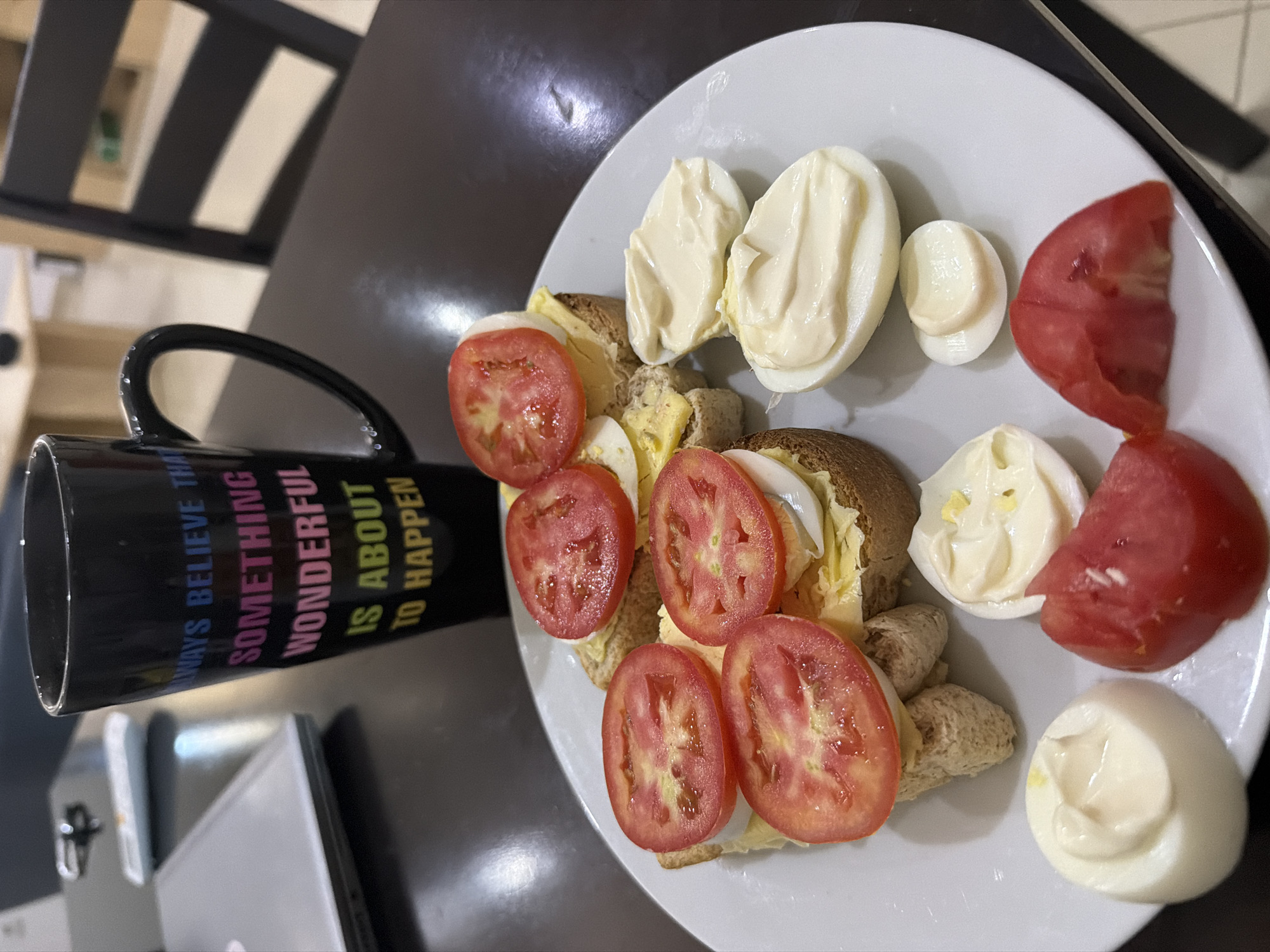The image, rotated counter-clockwise, displays a meal set on a dark table, likely indoors. A prominent white, circular plate is laden with food items, including several slices of red tomatoes, numerous hard-boiled eggs (some halved, some quartered), many of which are topped with a dollop of creamy white mayonnaise. Also on the plate are open-faced sandwich pieces made with brown bread, appearing to be layered with egg (possibly scrambled or egg salad) and some shredded green vegetable, topped with tomato slices.
To the left of the plate, a black ceramic mug rests on the table. The mug features colorful, motivational text that reads: "ALWAYS BELIEVE THAT SOMETHING WONDERFUL IS ABOUT TO HAPPEN". The background shows blurred dark furniture, possibly parts of chairs, and a hint of a light-colored object, which might be an electronic device, near the mug. No individuals are visible. The food suggests a breakfast or brunch meal.