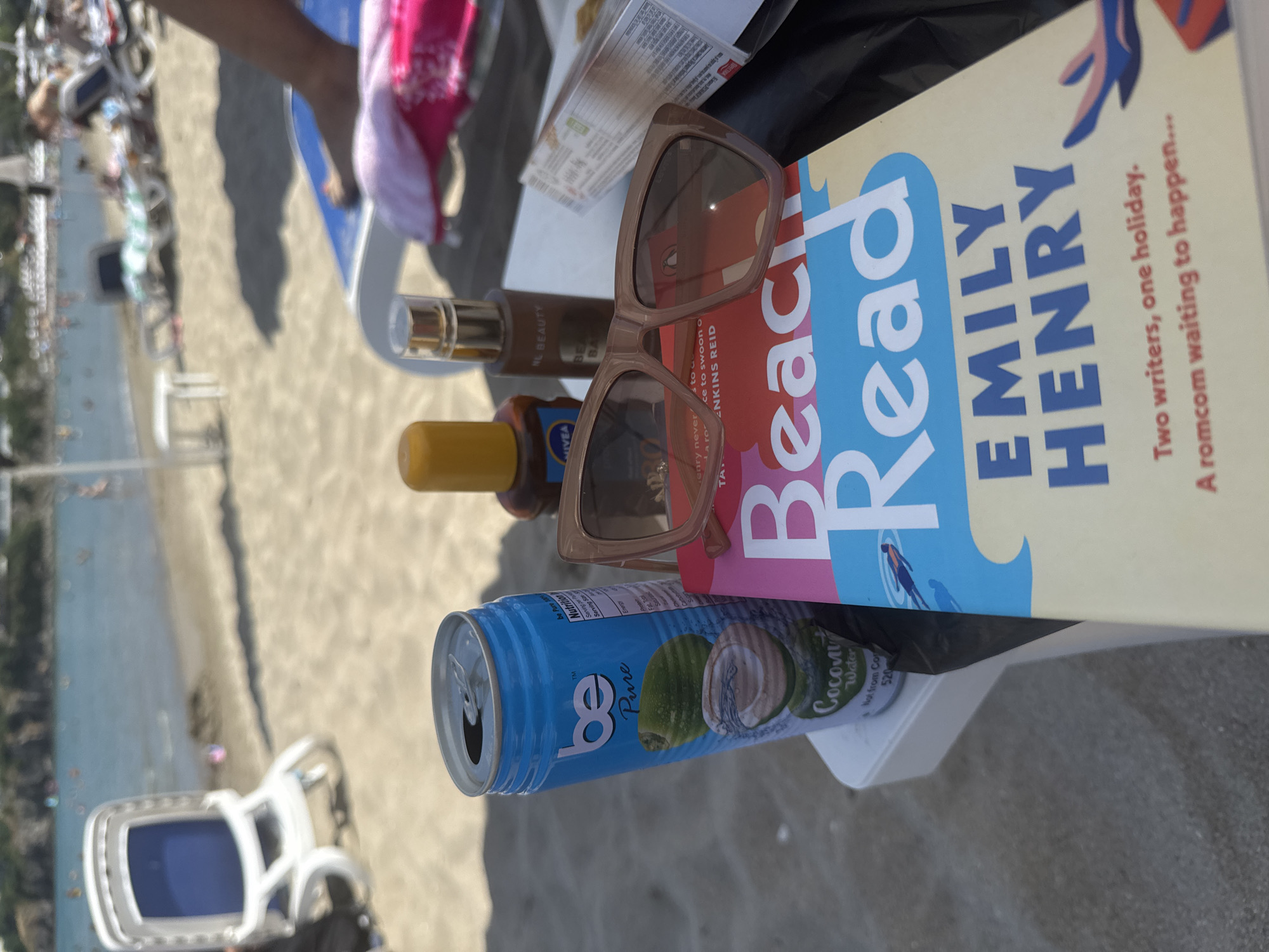 The image shows a close-up view of items placed on a white plastic table at a beach in Sozopol, Bulgaria. In the foreground, a blue can of "be Pure" coconut water is visible, next to a bottle of Nivea sun lotion and a bottle of beauty oil. Resting on top of a book titled "Beach Read" by Emily Henry are a pair of beige sunglasses. The book has a colorful cover with the title prominently displayed in large letters. In the background, the sandy beach is dotted with sun loungers and umbrellas. Several people are seen in the shallow water, enjoying the sea. The overall scene depicts a relaxed beach day.